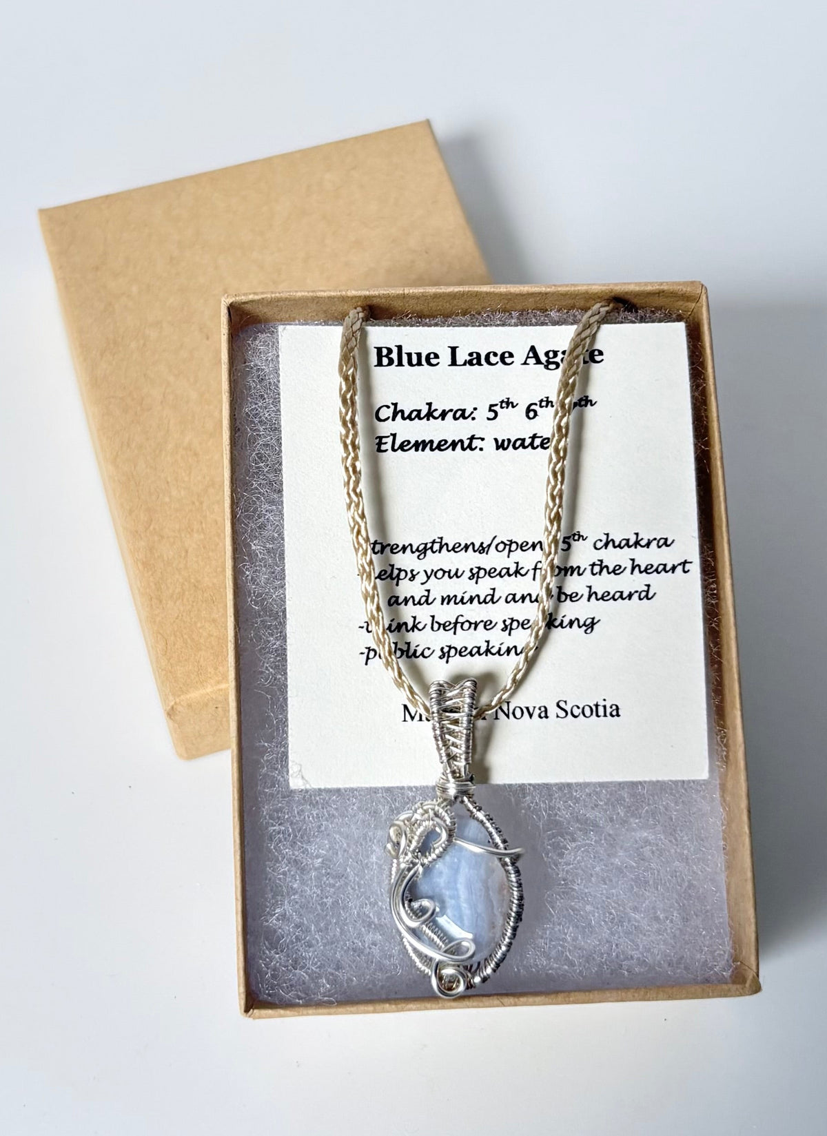 Necklace with a silver wire wrapped blue lace agate pendant on an information card with text explaining the folklore and metaphysical properties placed in a brown paper kraft jewelry box for gift giving all on a white surface.