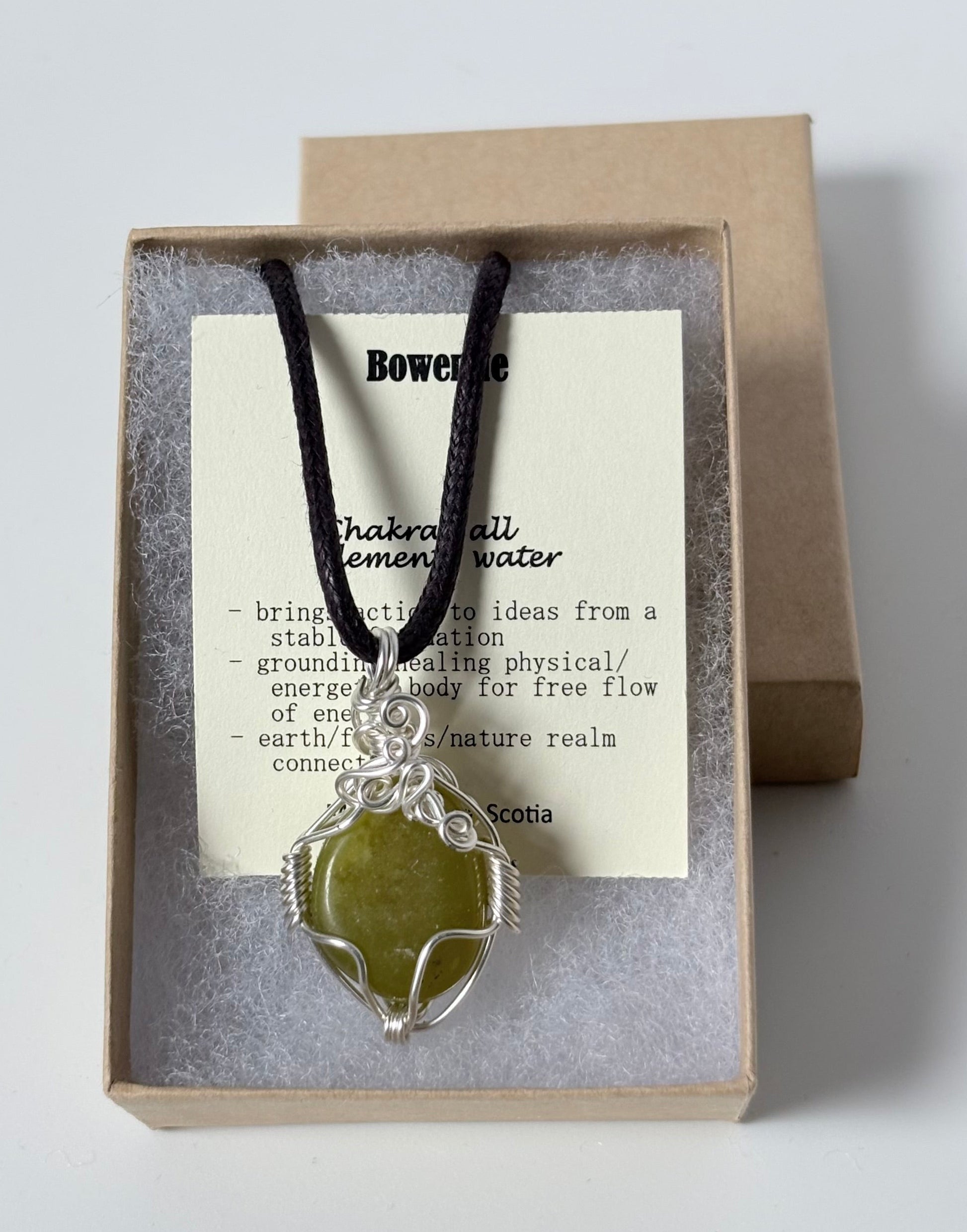 Necklace with a silver wire wrapped bowenite pendant on an information card with text explaining the folklore and metaphysical properties placed in a brown paper kraft jewelry box for gift giving all on a white surface.