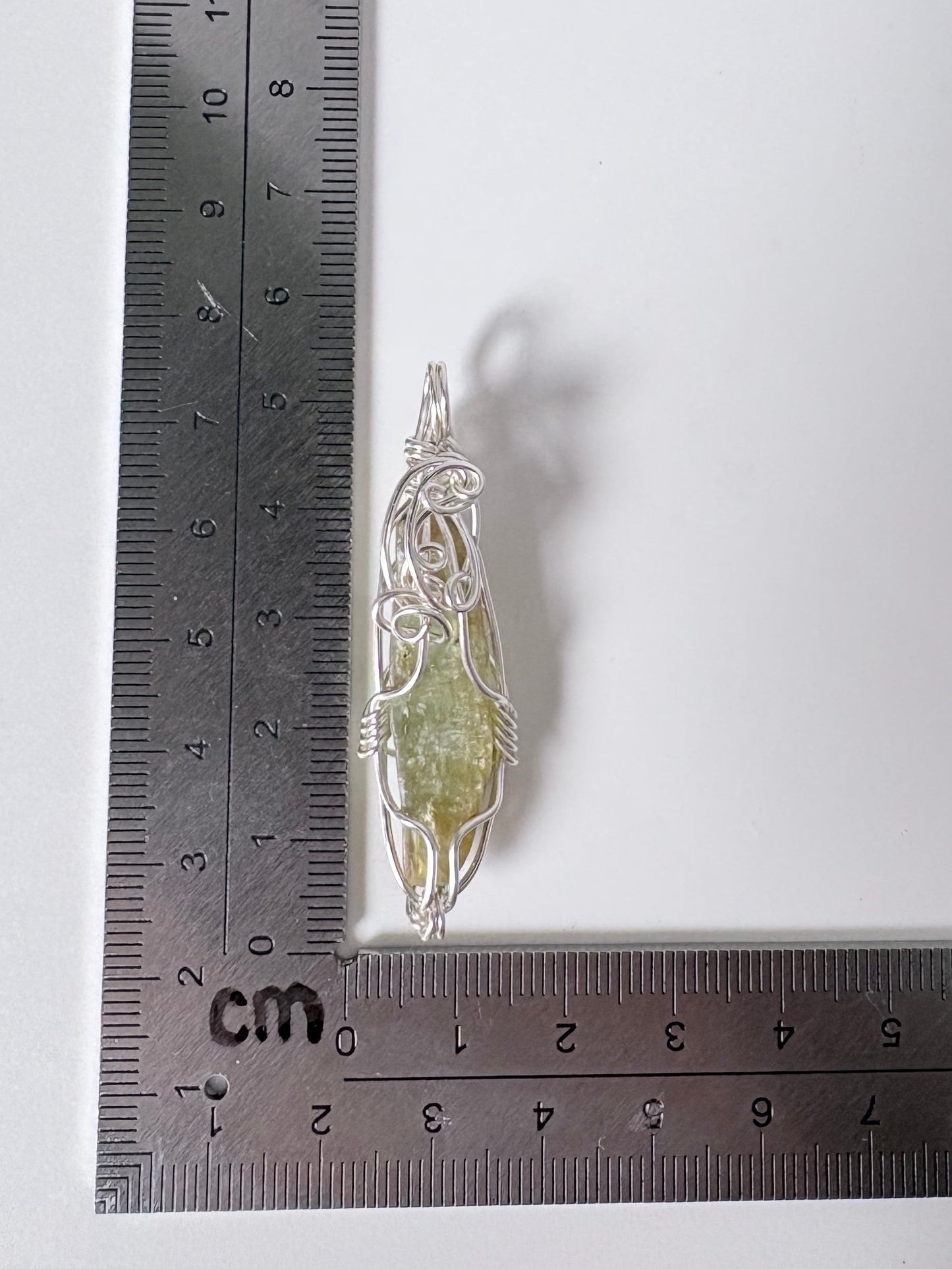 silver wire wrapped green kyanite pendant on a white background with a ruler for scale.