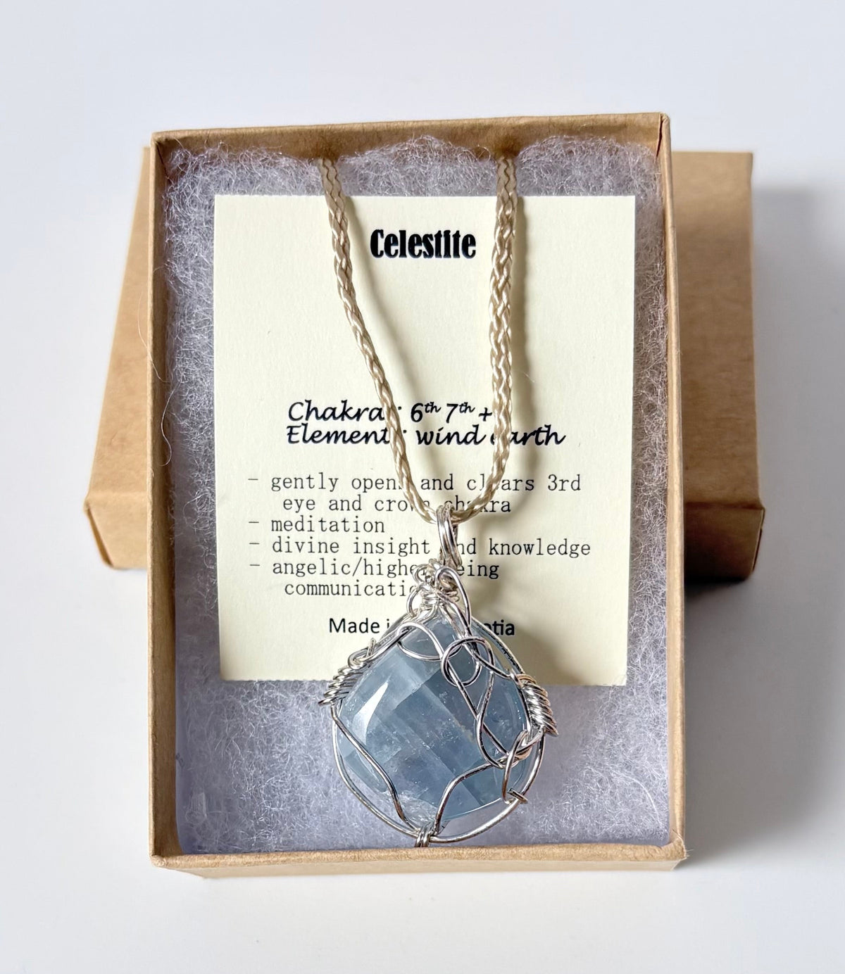 Necklace with a silver wire wrapped celestite pendant on an information card with text explaining the folklore and metaphysical properties placed in a brown paper kraft jewelry box for gift giving all on a white surface.