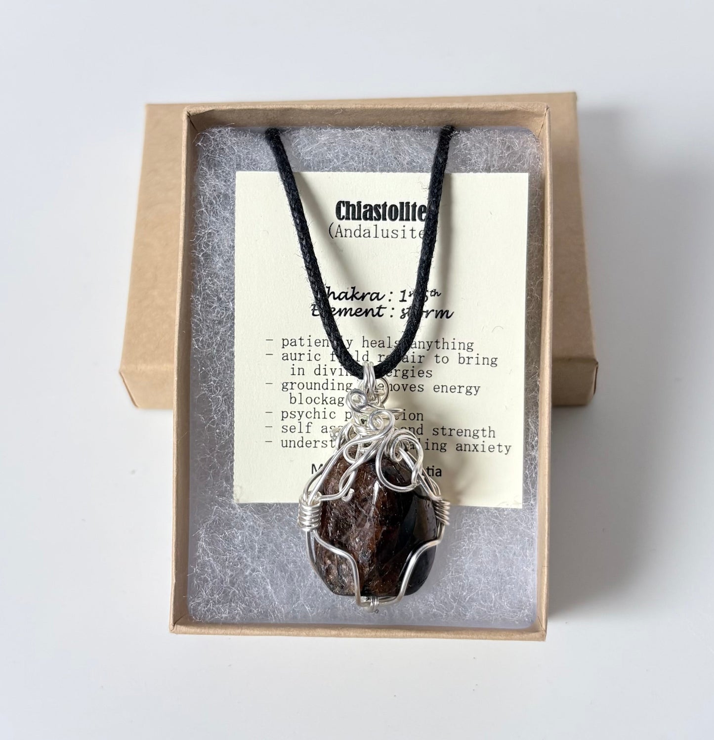 Necklace with a silver wire wrapped chiastolite pendant on an information card with text explaining the folklore and metaphysical properties placed in a brown paper kraft jewelry box for gift giving all on a white surface.