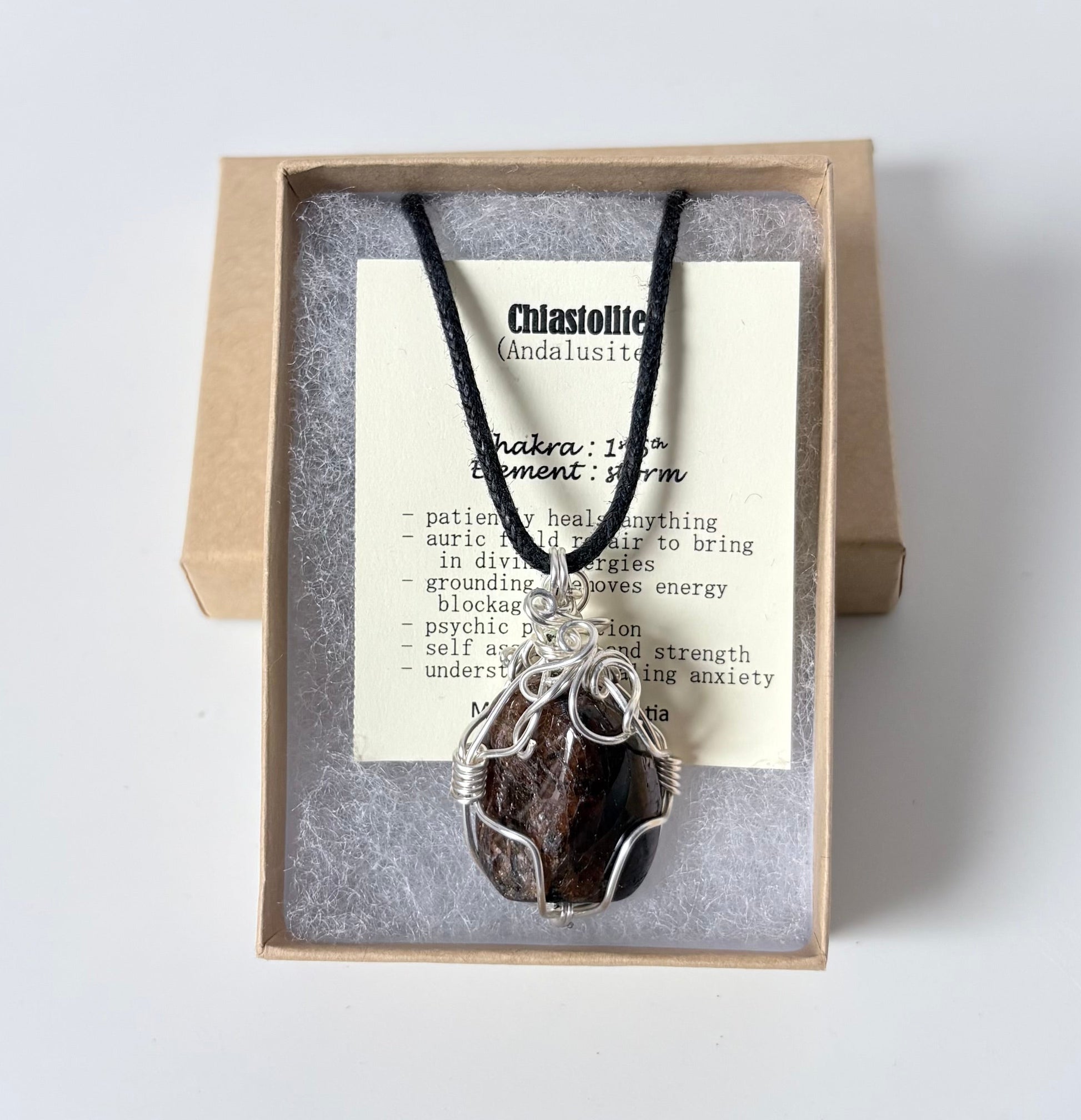 Necklace with a silver wire wrapped chiastolite pendant on an information card with text explaining the folklore and metaphysical properties placed in a brown paper kraft jewelry box for gift giving all on a white surface.