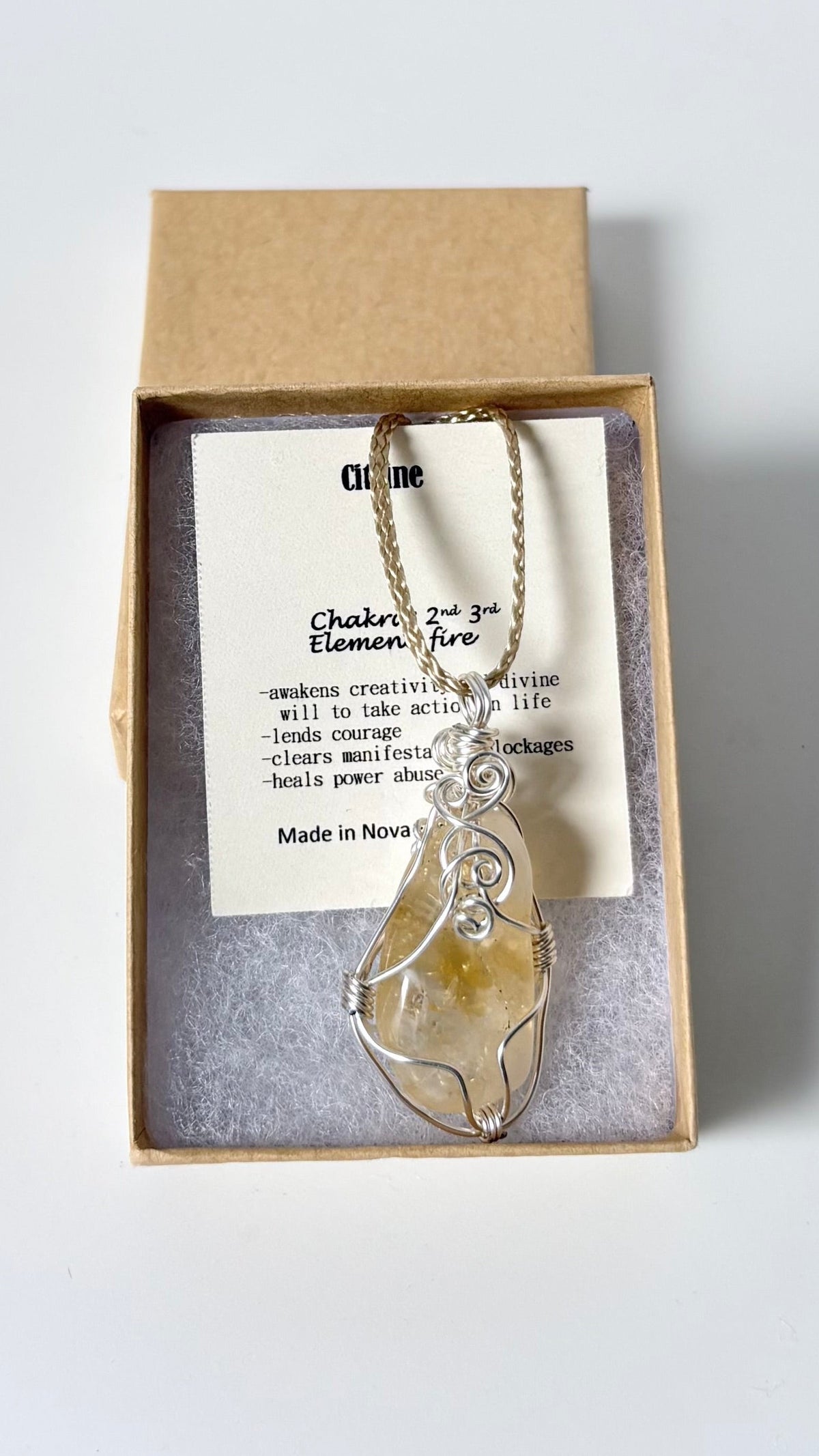 Necklace with a silver wire wrapped citrine pendant on an information card with text explaining the folklore and metaphysical properties placed in a brown paper kraft jewelry box for gift giving all on a white surface.