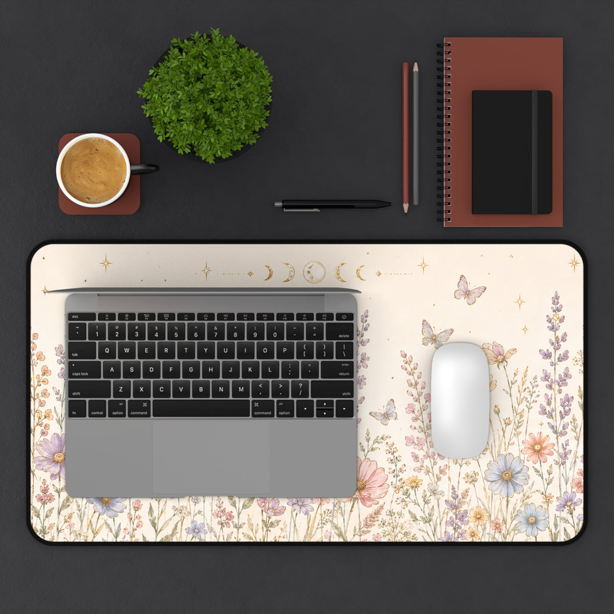 12 by 22 inch botanical desk mat shown on a dark desktop with a laptop and white mouse for size reference. The cream-colored keyboard and mouse pad features pastel wildflowers, butterflies, gold stars and moon phases, with rounded corners and a black stitched border. A cup of coffee, green plant, pens, pencils, spiral notebook and small black journal are arranged around it to show the mat’s scale in a workspace.