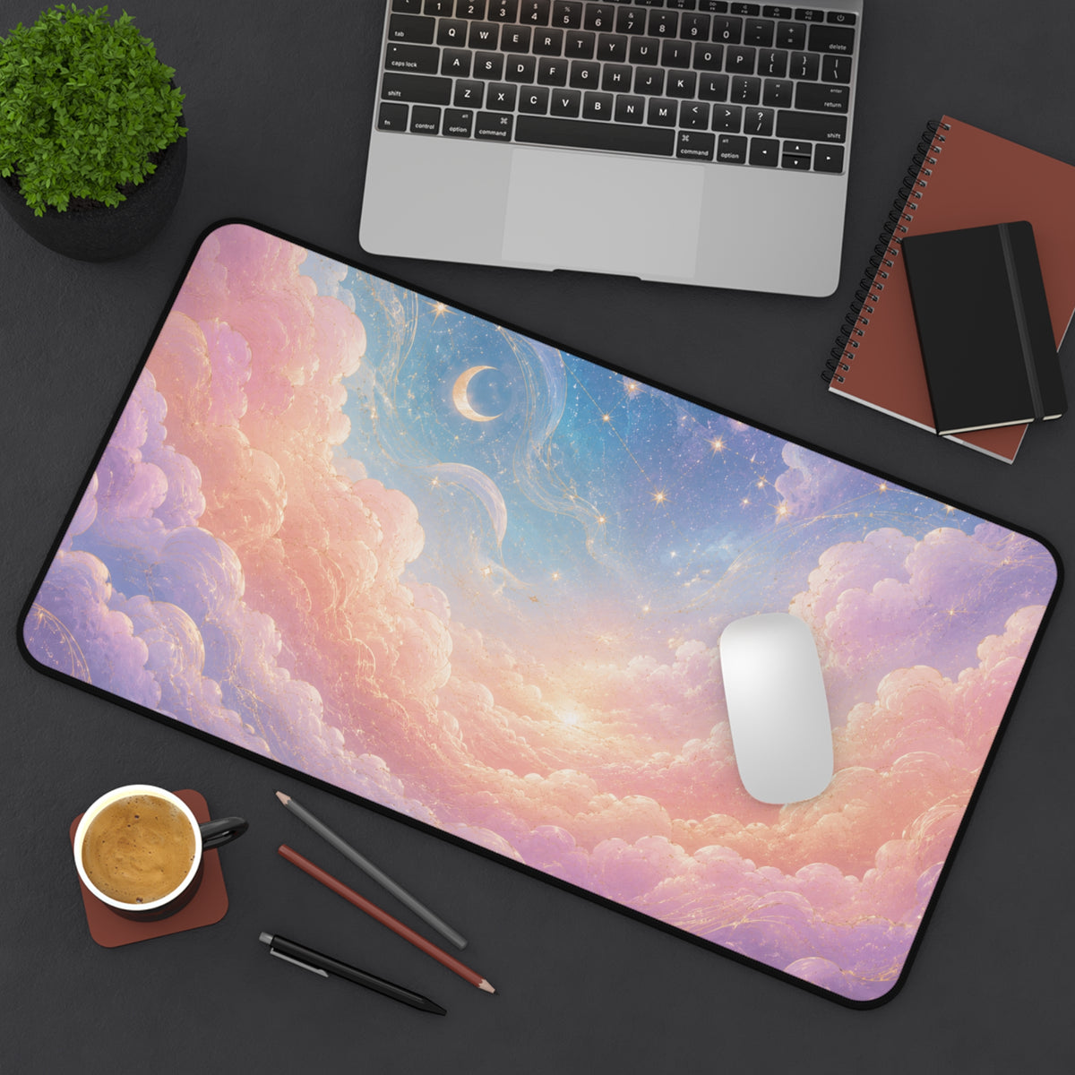 12 by 22 inch pastel celestial desk mat shown on a dark desktop with a white mouse, laptop, coffee cup, plant, pencils, pen, spiral notebook and small black journal for size reference. The large keyboard and mouse pad features dreamy pink and lavender clouds, a blue starry sky, crescent moon, glowing stars and gold swirl details, with rounded corners and a black stitched border.