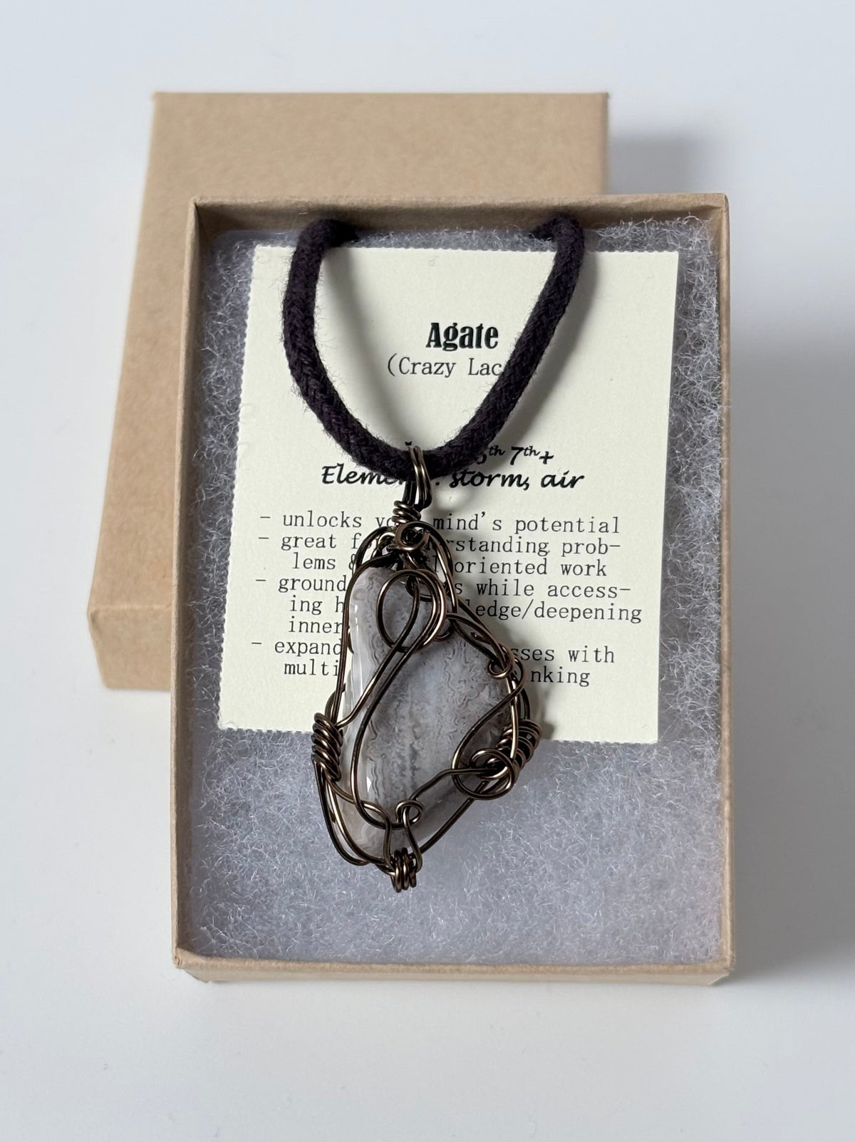 Necklace with an antique brass wire wrapped crazy lace agate pendant on an information card with text explaining the folklore and metaphysical properties placed in a brown paper kraft jewelry box for gift giving all on a white surface.