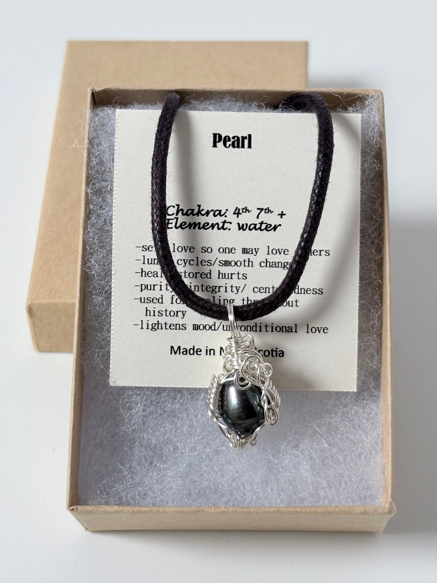 Necklace with a silver wire wrapped dark pearl pendant on an information card with text explaining the folklore and metaphysical properties placed in a brown paper kraft jewelry box for gift giving all on a white surface.
