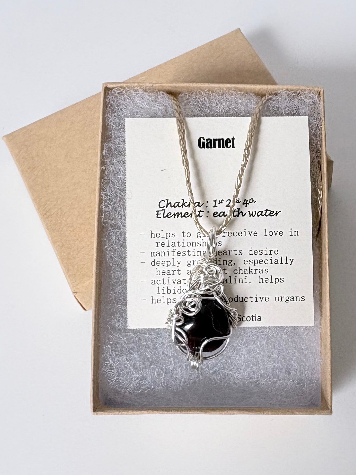 Necklace with a silver wire wrapped garnet pendant on an information card with text explaining the folklore and metaphysical properties placed in a brown paper kraft jewelry box for gift giving all on a white surface.