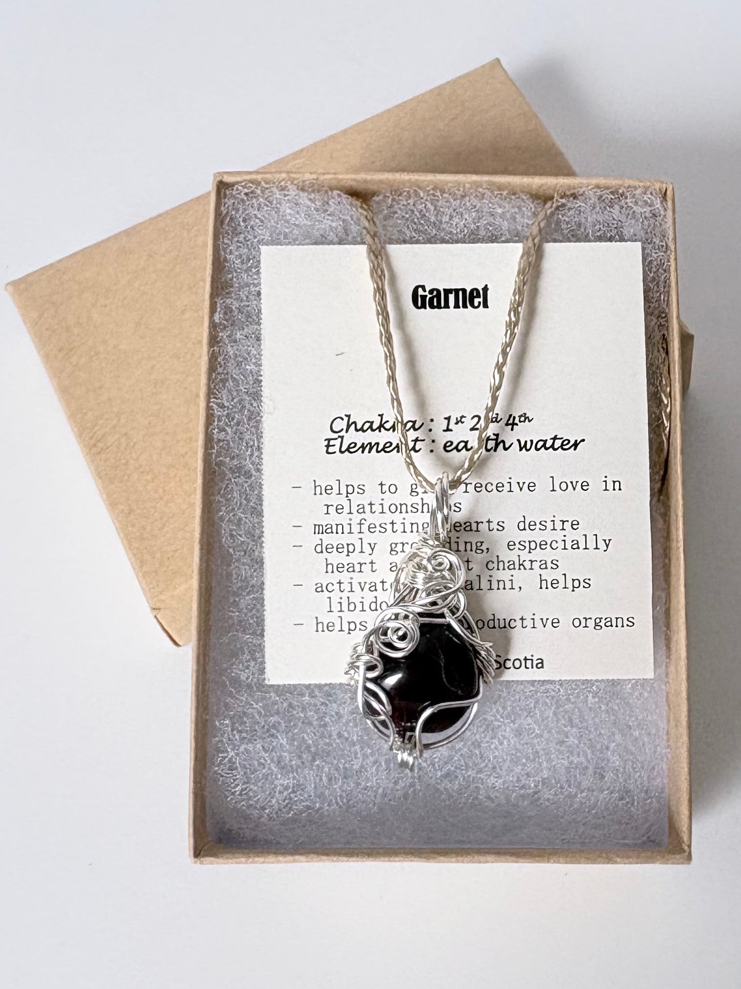Necklace with a silver wire wrapped garnet pendant on an information card with text explaining the folklore and metaphysical properties placed in a brown paper kraft jewelry box for gift giving all on a white surface.