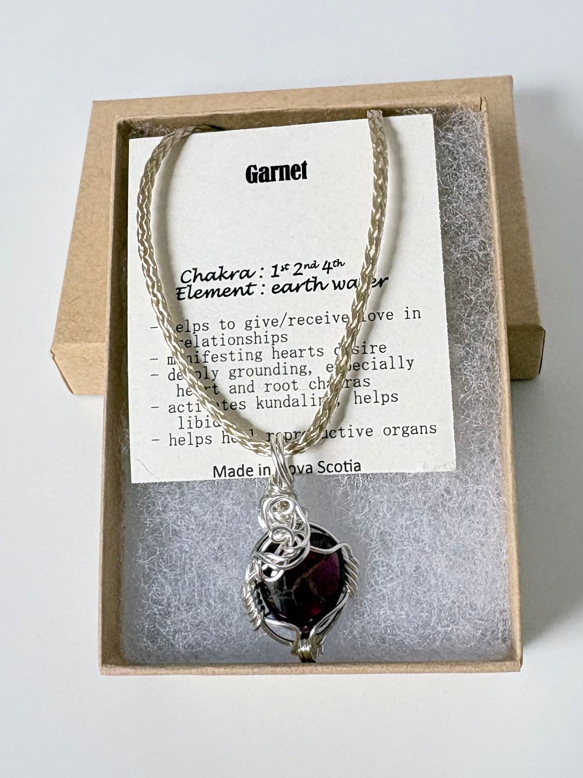 Necklace with a silver wire wrapped garnet pendant on an information card with text explaining the folklore and metaphysical properties placed in a brown paper kraft jewelry box for gift giving all on a white surface.