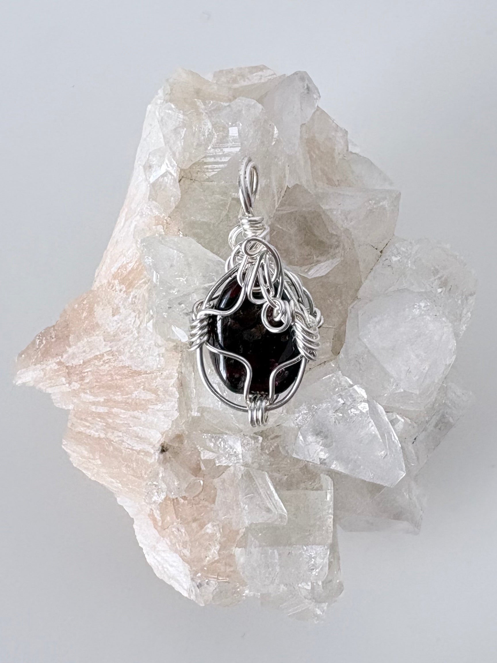 silver wire wrapped garnet pendant displayed on a clear apophyllite cluster all on a white surface.