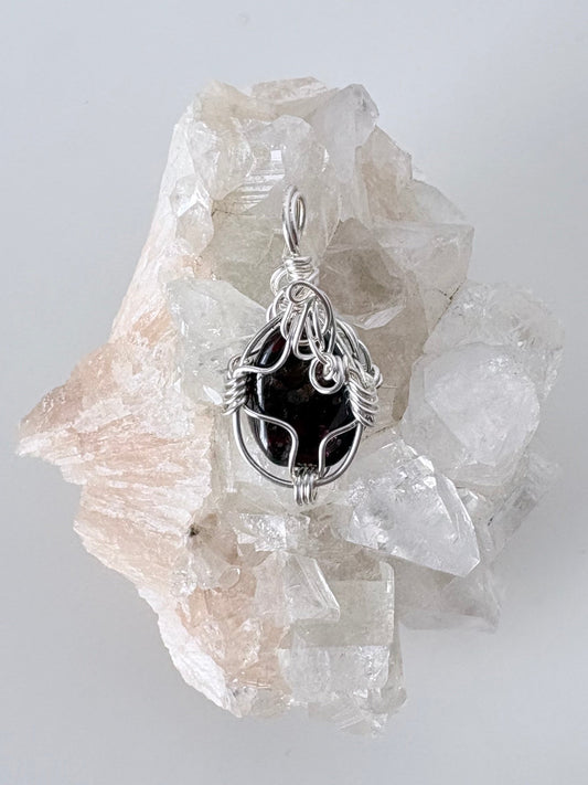 silver wire wrapped garnet pendant displayed on a clear apophyllite cluster all on a white surface.