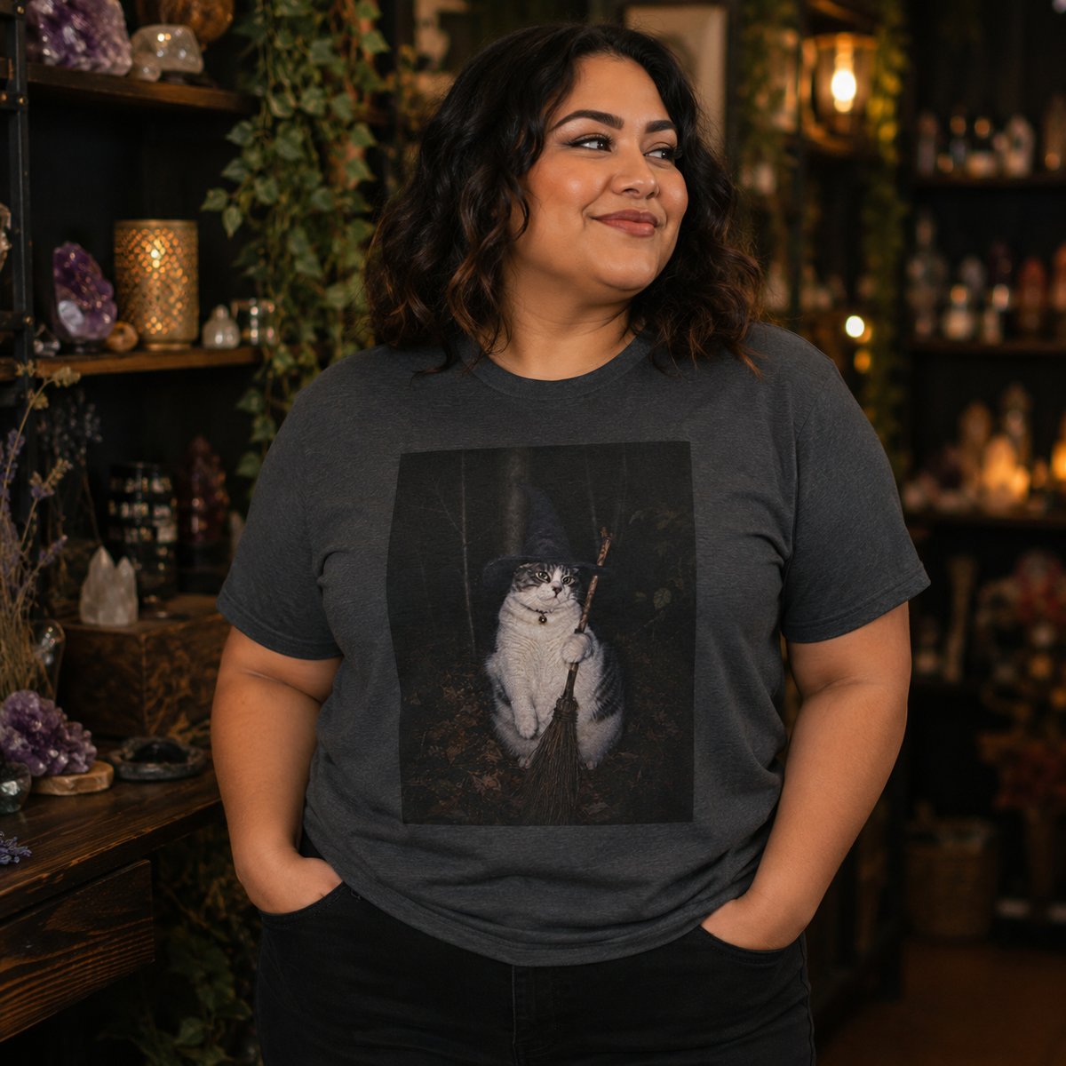 Model wearing a dark heather grey graphic t-shirt featuring a photo of a cat wearing a black witch hat and a collar with a round bell, holding a broom in a dark woodland setting. the model is in a cozy mystical shop with crystals, plants and warm lighting.