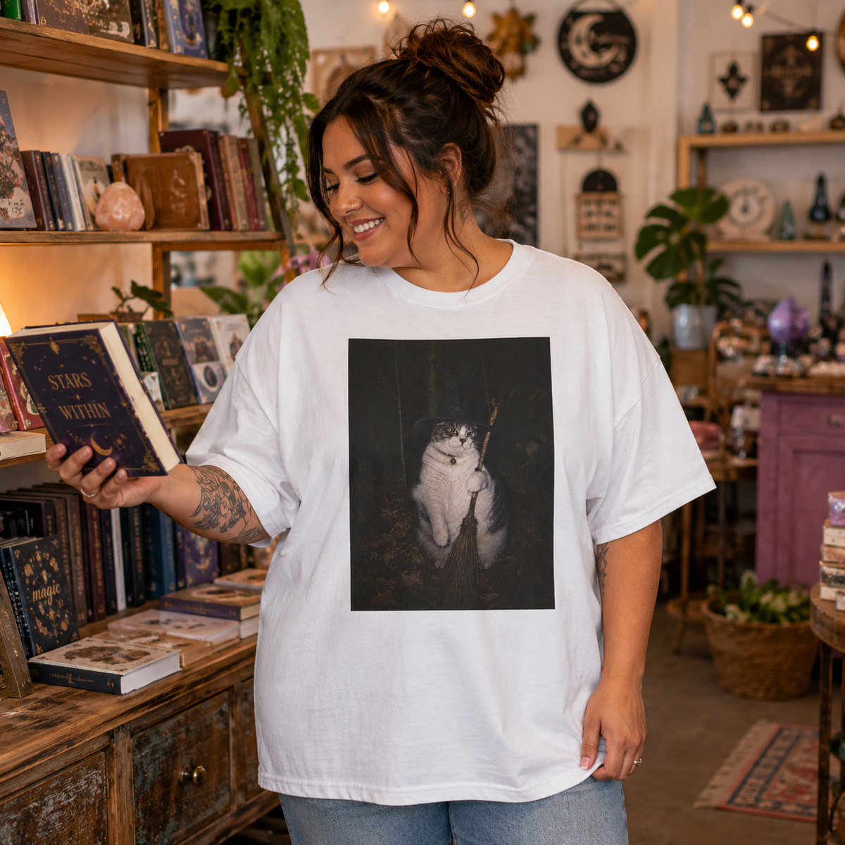Model wearing an oversized white graphic t-shirt featuring a photo of a cat wearing a black witch hat and a collar with a round bell, holding a broom in a dark woodland setting. the model is in a cozy spiritual shop with books, plants and mystical decor.