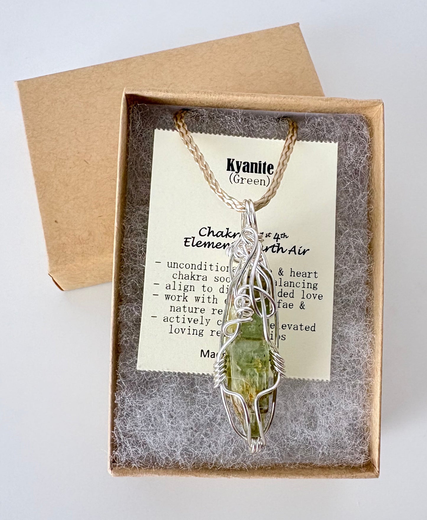 Necklace with a silver wire wrapped kyanite (green) pendant on an information card with text explaining the folklore and metaphysical properties placed in a brown paper kraft jewelry box for gift giving all on a white surface.