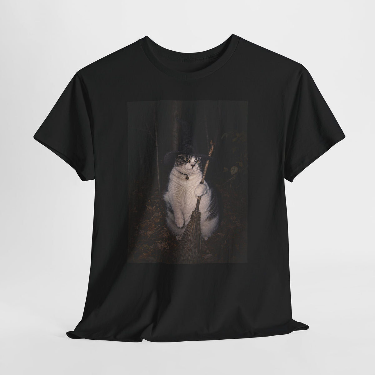 Front of a black graphic t-shirt featuring a photo of a cat wearing a black witch hat and a collar with a round bell, holding a broom in a dark woodland setting. The t-shirt is shown on a white background.