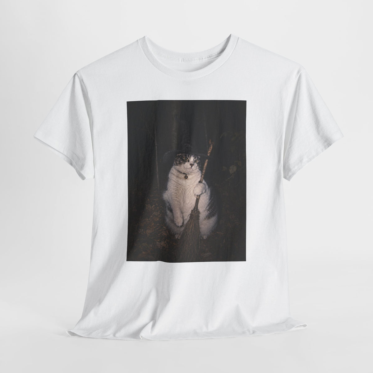 Front of a white graphic t-shirt featuring a photo of a cat wearing a black witch hat and a collar with a round bell, holding a broom in a dark woodland setting. The t-shirt is shown on a white background.