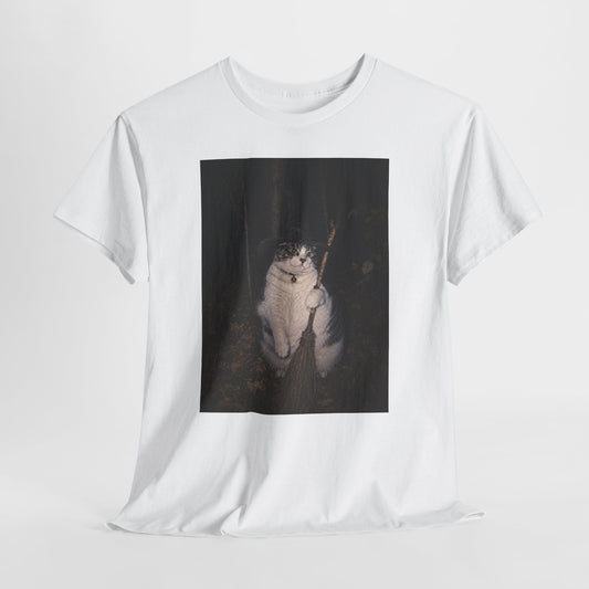 Front of a white graphic t-shirt featuring a photo of a cat wearing a black witch hat and a collar with a round bell, holding a broom in a dark woodland setting. The t-shirt is shown on a white background.