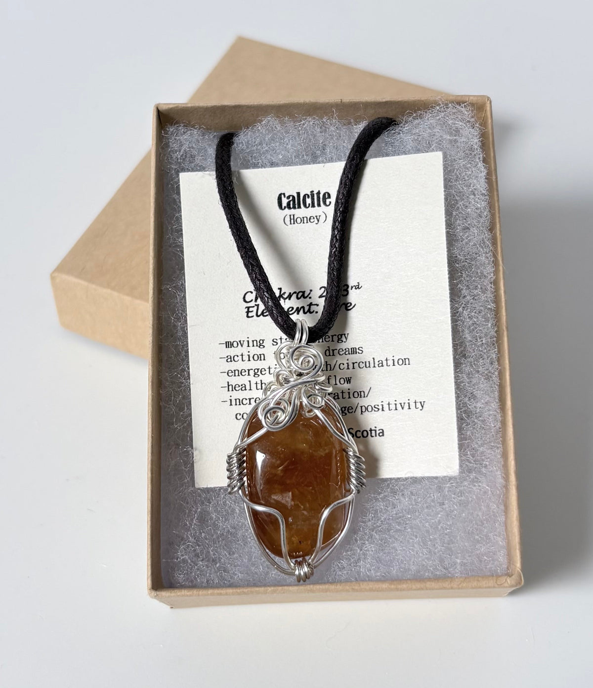Necklace with a silver wire wrapped calcite (honey) pendant on an information card with text explaining the folklore and metaphysical properties placed in a brown paper kraft jewelry box for gift giving all on a white surface.