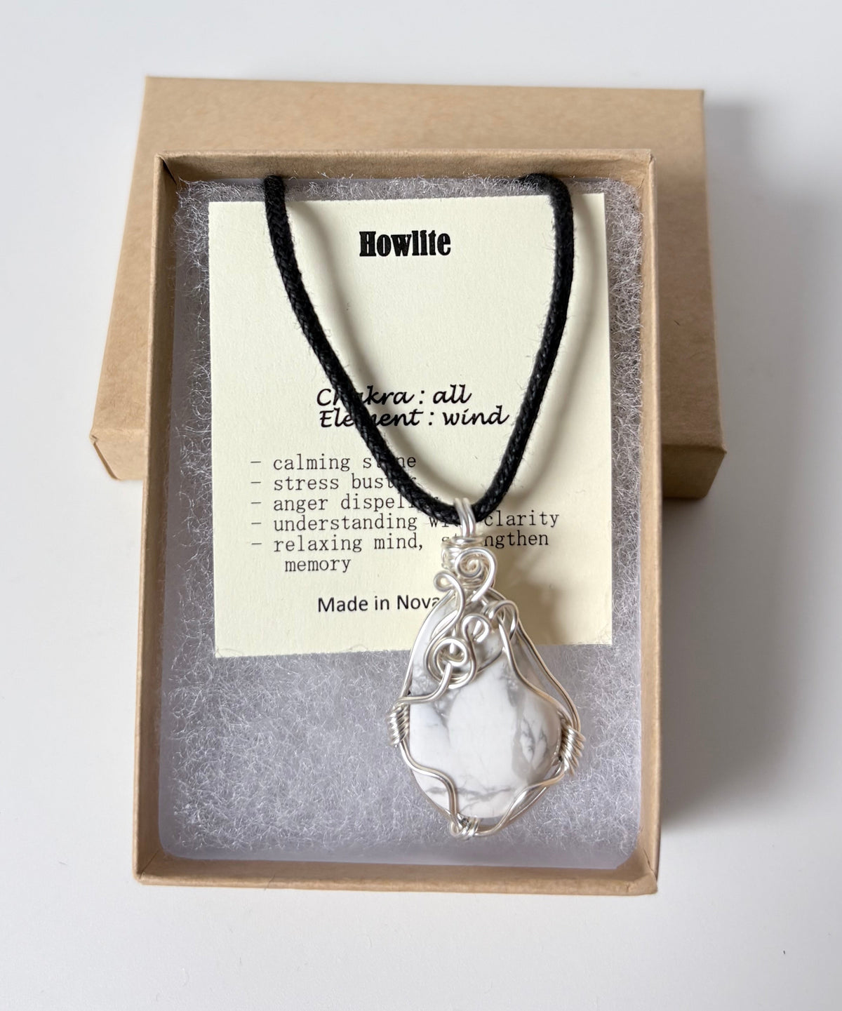 Necklace with a silver wire wrapped howlite pendant on an information card with text explaining the folklore and metaphysical properties placed in a brown paper kraft jewelry box for gift giving all on a white surface.