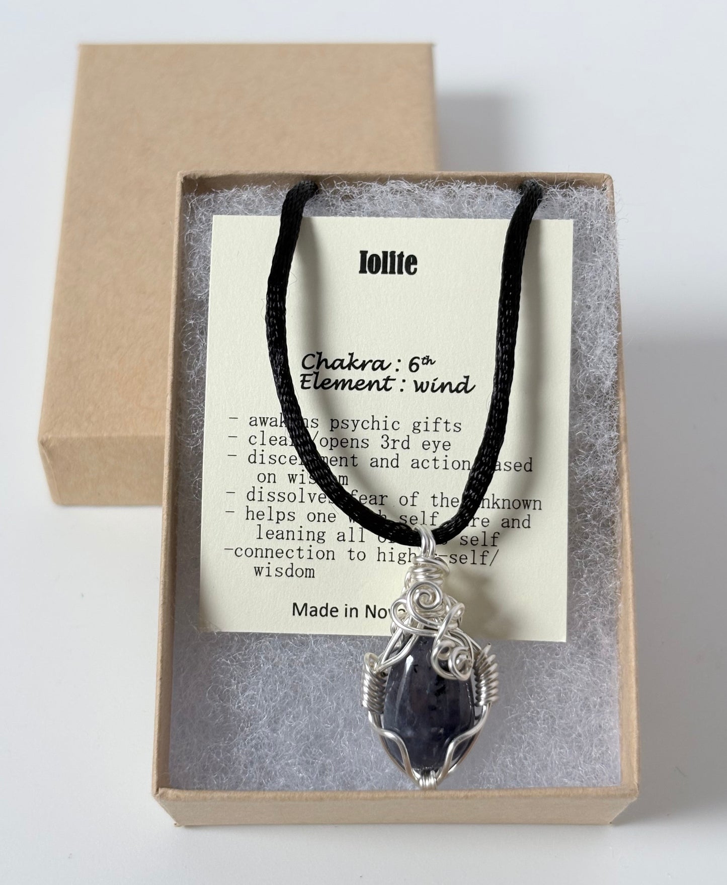 Necklace with a silver wire wrapped iolite pendant on an information card with text explaining the folklore and metaphysical properties placed in a brown paper kraft jewelry box for gift giving all on a white surface.