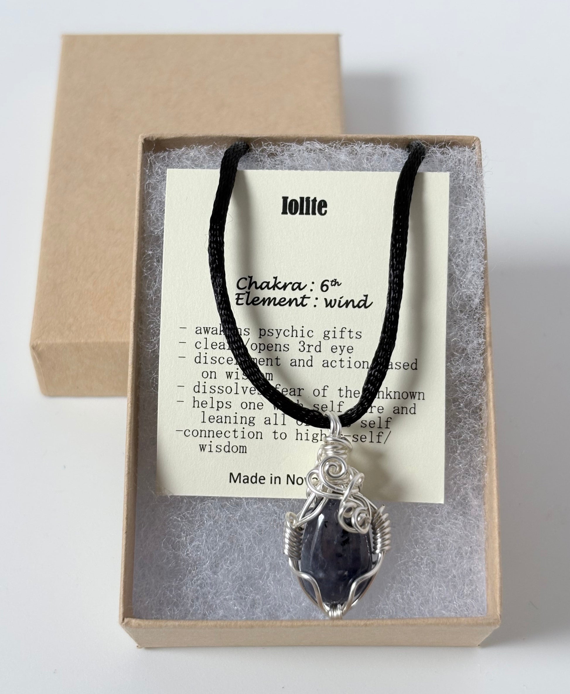 Necklace with a silver wire wrapped iolite pendant on an information card with text explaining the folklore and metaphysical properties placed in a brown paper kraft jewelry box for gift giving all on a white surface.