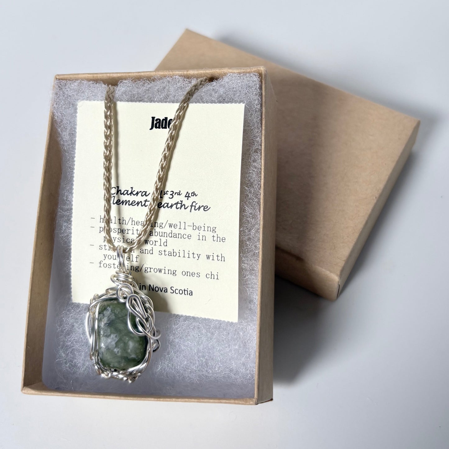 Necklace with a silver wire wrapped jade pendant on an information card with text explaining the folklore and metaphysical properties placed in a brown paper kraft jewelry box for gift giving all on a white surface.