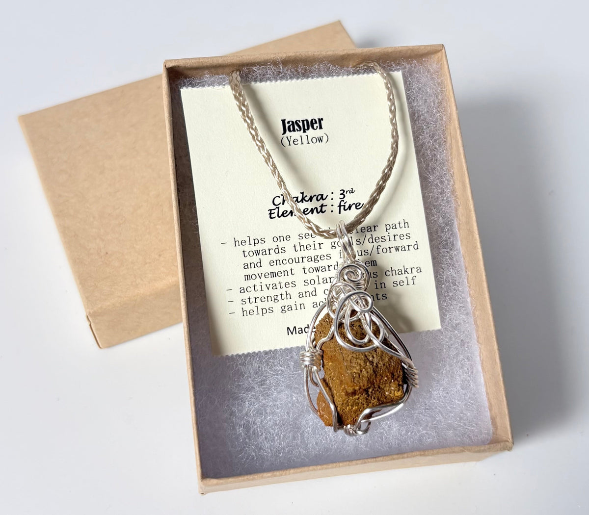 Necklace with a silver wire wrapped jasper (yellow) pendant on an information card with text explaining the folklore and metaphysical properties placed in a brown paper kraft jewelry box for gift giving all on a white surface.