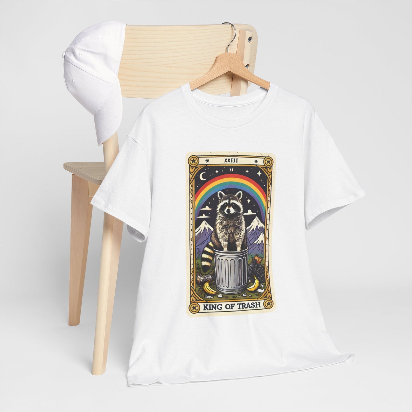White t-shirt with a graphic design of a raccoon on a trash can with a rainbow, hanging on a wooden hanger against a white background.