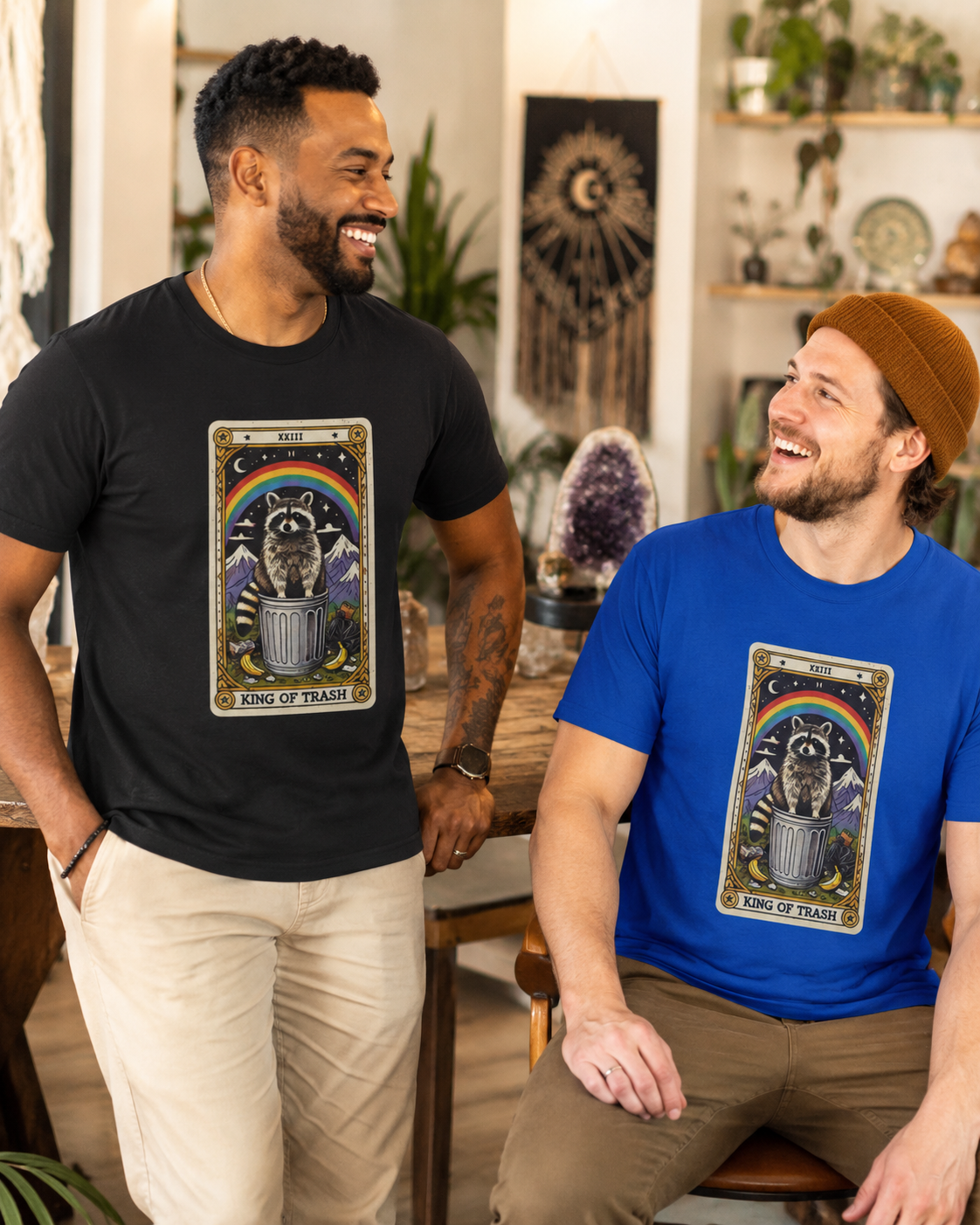 Two men wearing t-shirts with a tarot card-style design featuring the king of trash tarot card that has a raccoon  standing on a trash can under a rainbow. One shirt is black, the other shirt is royal blue.