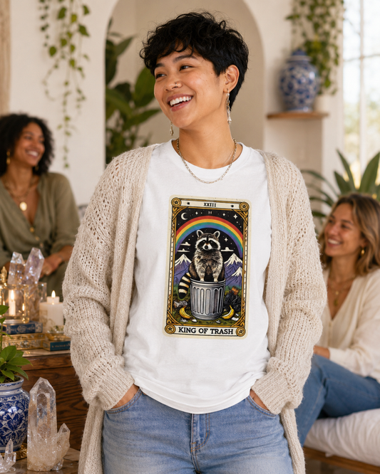 Person wearing a white t-shirt with a graphic design of a raccoon in a trash can and rainbow, standing in a room with plants and people.