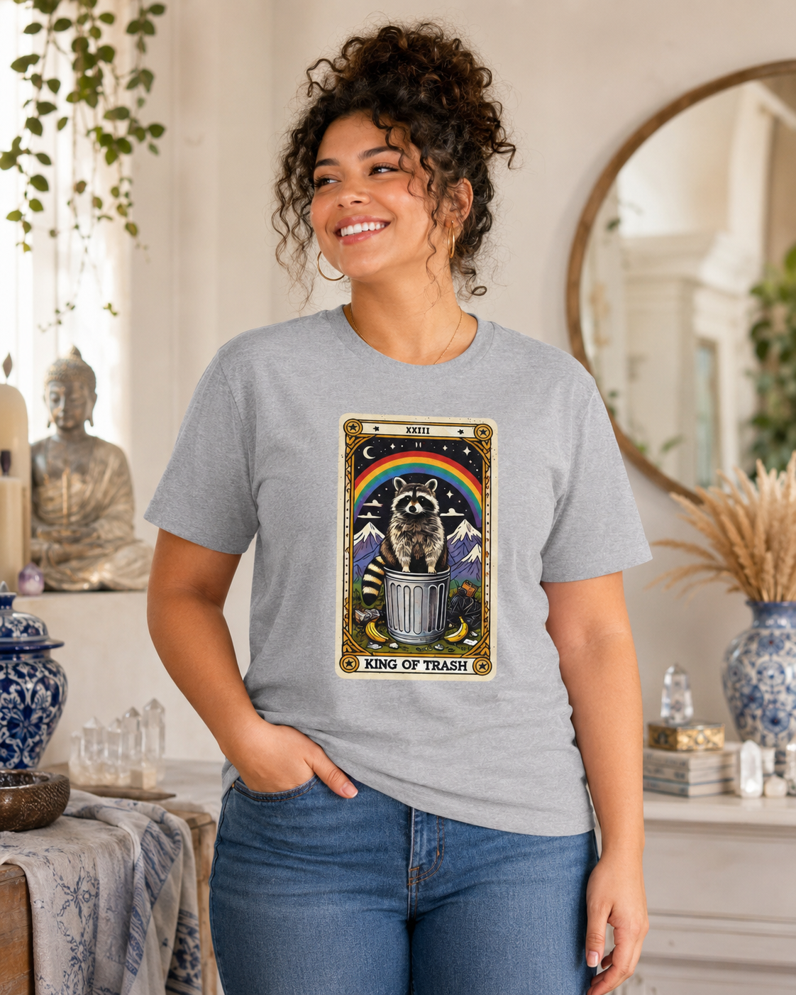 A model wearing a heather grey t-shirt with a tarot card-style design featuring the king of trash tarot card that has a raccoon standing on a trash can under a rainbow. The card has a roman numeral on the top and "King Of Trash" written on the bottom of the card image.