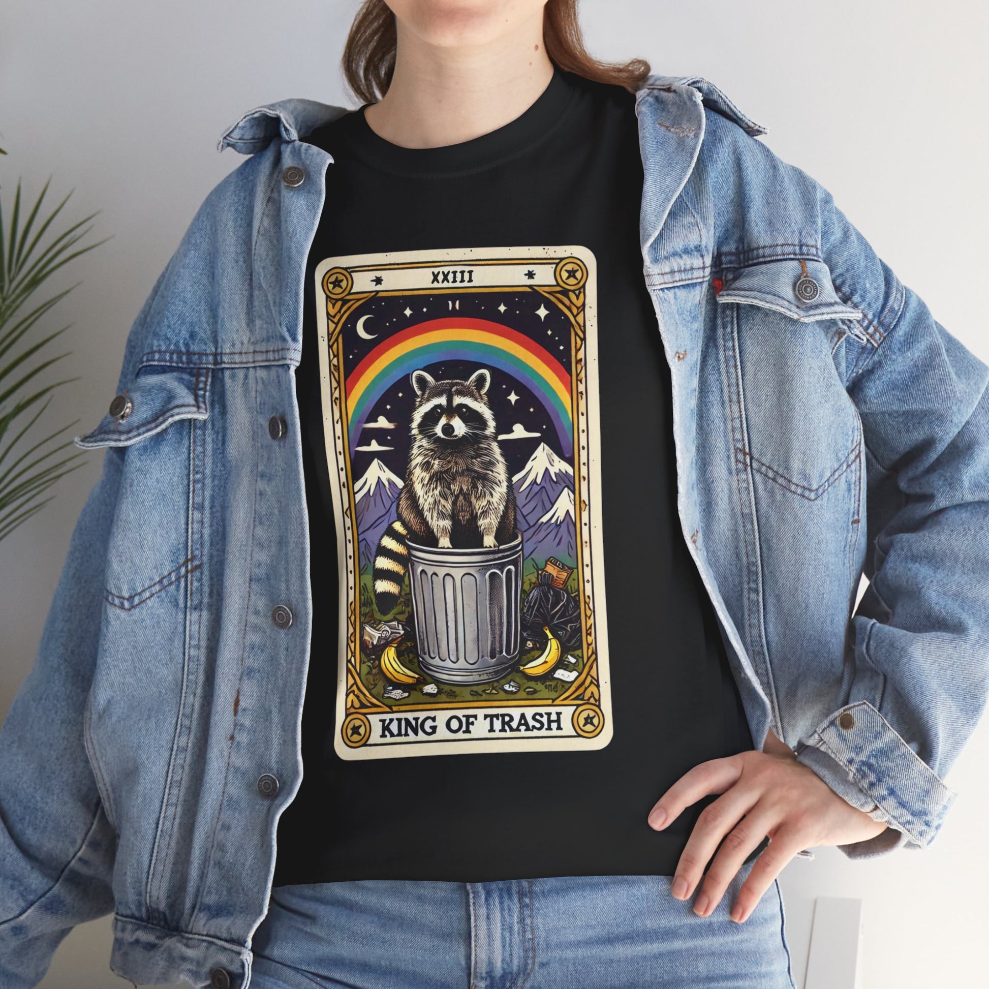 Model wearing a black t-shirt with a tarot card-style design featuring the king of trash tarot card that has a raccoon standing on a trash can under a rainbow. The card has a roman numeral on the top and "King Of Trash" written on the bottom of the card image. They are wearing a blue jean jacket over the T-shirt.