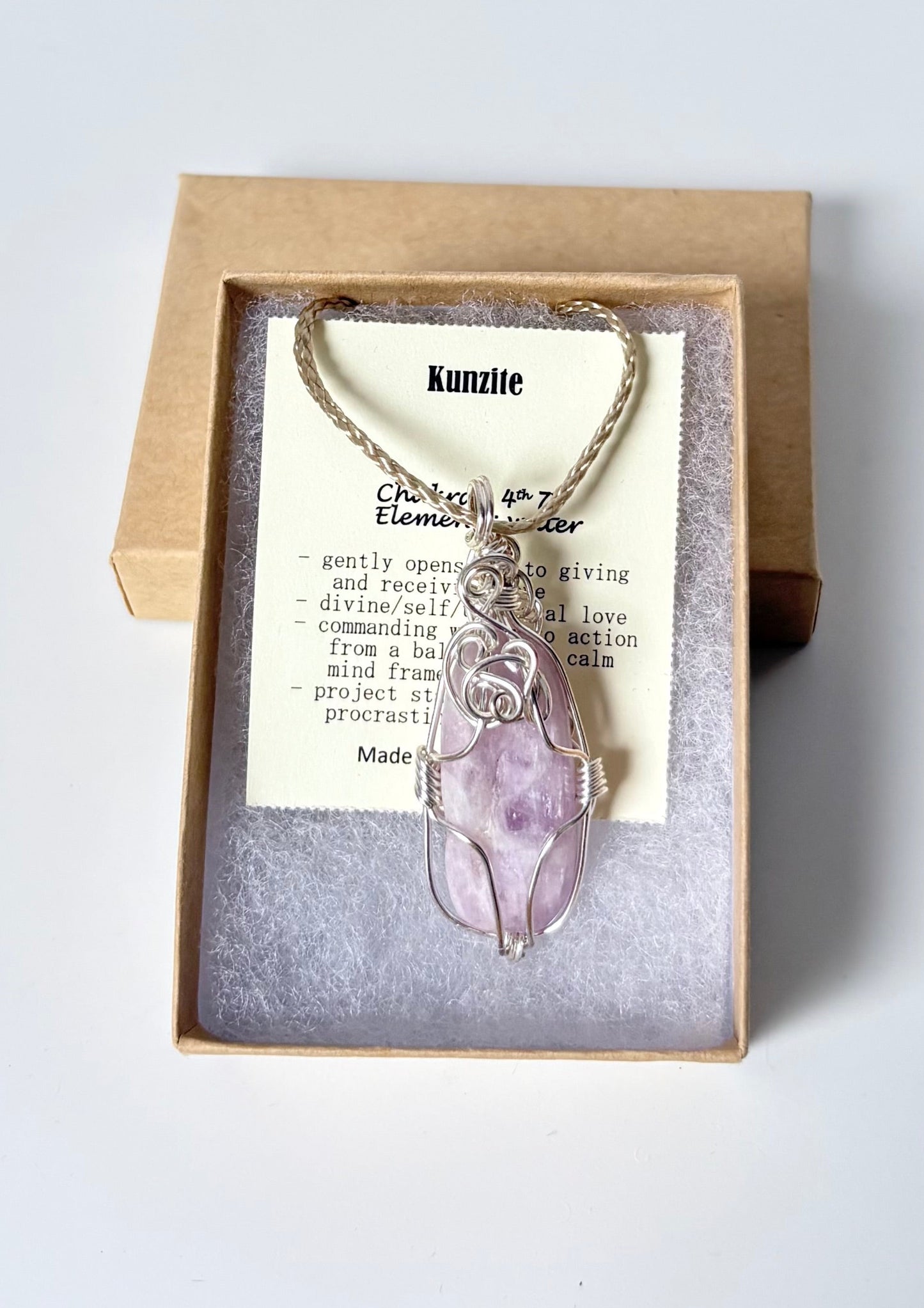 Necklace with a silver wire wrapped Kunzite pendant on an information card with text explaining the folklore and metaphysical properties placed in a brown paper kraft jewelry box for gift giving all on a white surface.
