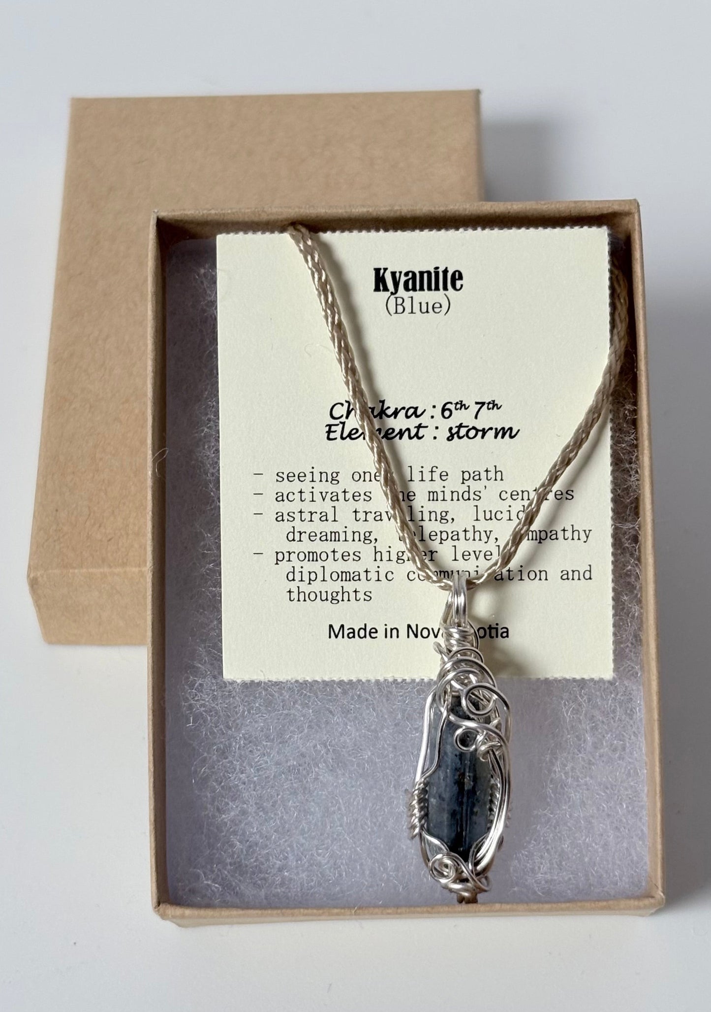 Necklace with a silver wire wrapped kyanite (blue) pendant on an information card with text explaining the folklore and metaphysical properties placed in a brown paper kraft jewelry box for gift giving all on a white surface.