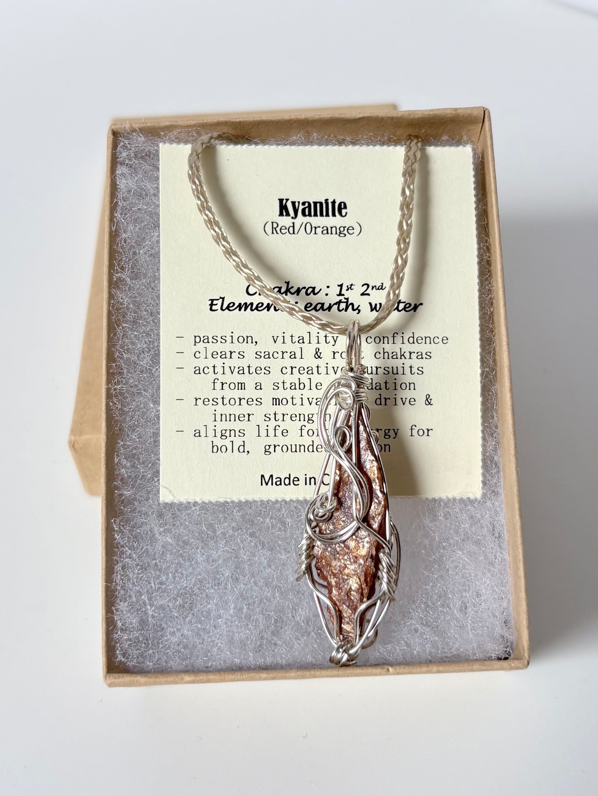 Necklace with a silver wire wrapped kyanite (red/orange) pendant on an information card with text explaining the folklore and metaphysical properties placed in a brown paper kraft jewelry box for gift giving all on a white surface.
