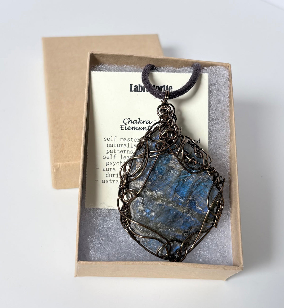 Necklace with an antique brass wire wrapped labradorite pendant on an information card with text explaining the folklore and metaphysical properties placed in a brown paper kraft jewelry box for gift giving all on a white surface.