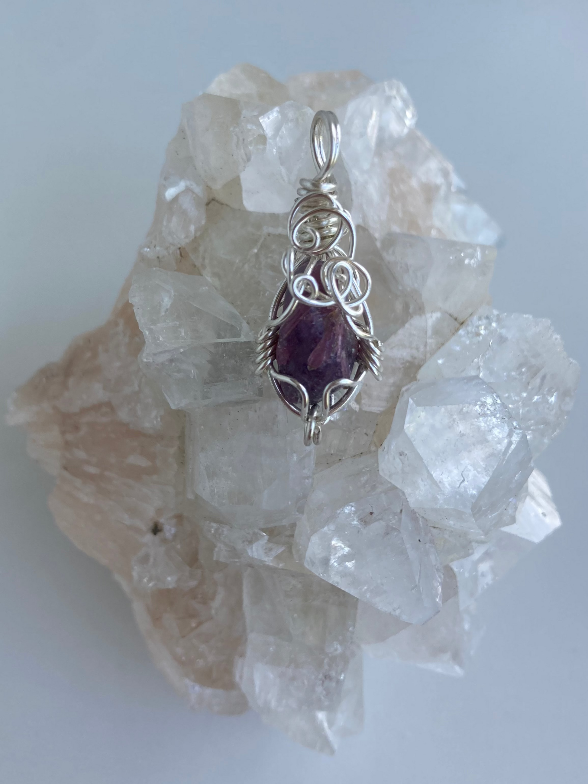 Lepidolite Pendant – Back view of a wire-wrapped purple lepidolite stone pendant, handcrafted in silver-plated copper wire with a unique spiral design — photographed on a clear apophyllite crystal cluster surface.