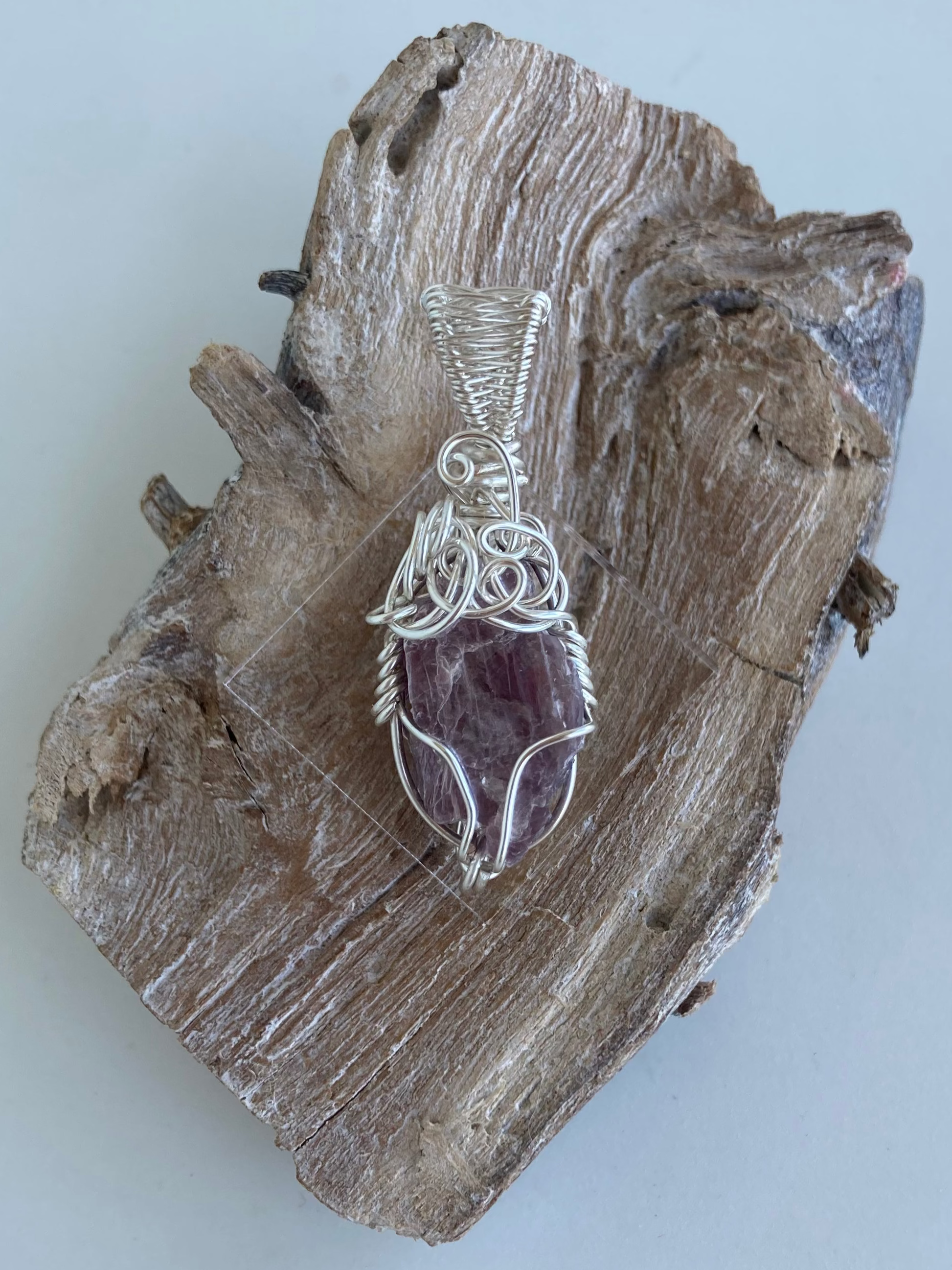 Lepidolite Pendant – Back view of a wire-wrapped purple lepidolite stone pendant, handcrafted in silver-plated copper wire with a unique spiral design — photographed on a wood surface with a white background.