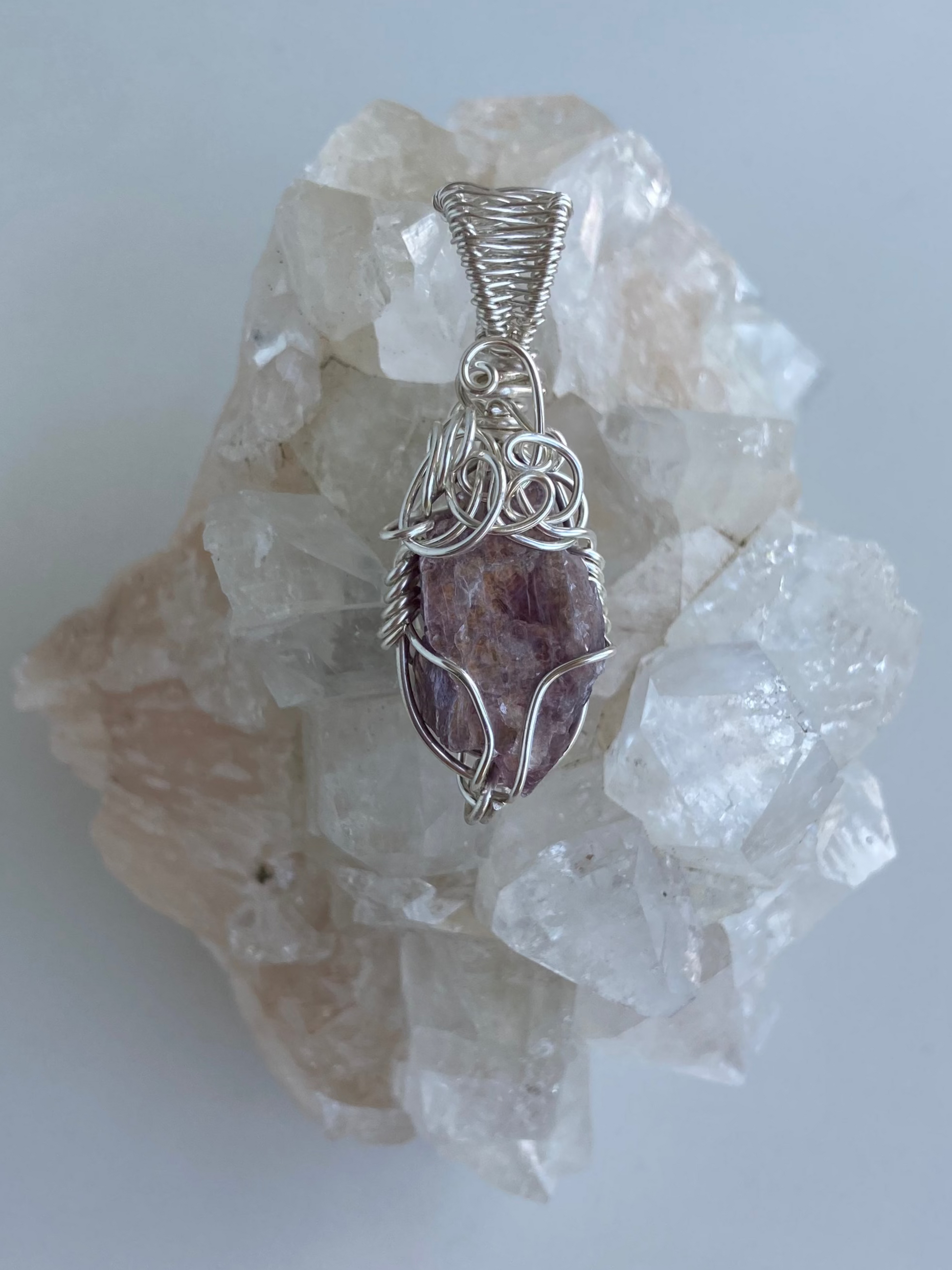 Lepidolite Pendant – Back view of a wire-wrapped purple lepidolite stone pendant, handcrafted in silver-plated copper wire with a unique spiral design — photographed on a clear apophyllite crystal cluster surface with a white background.