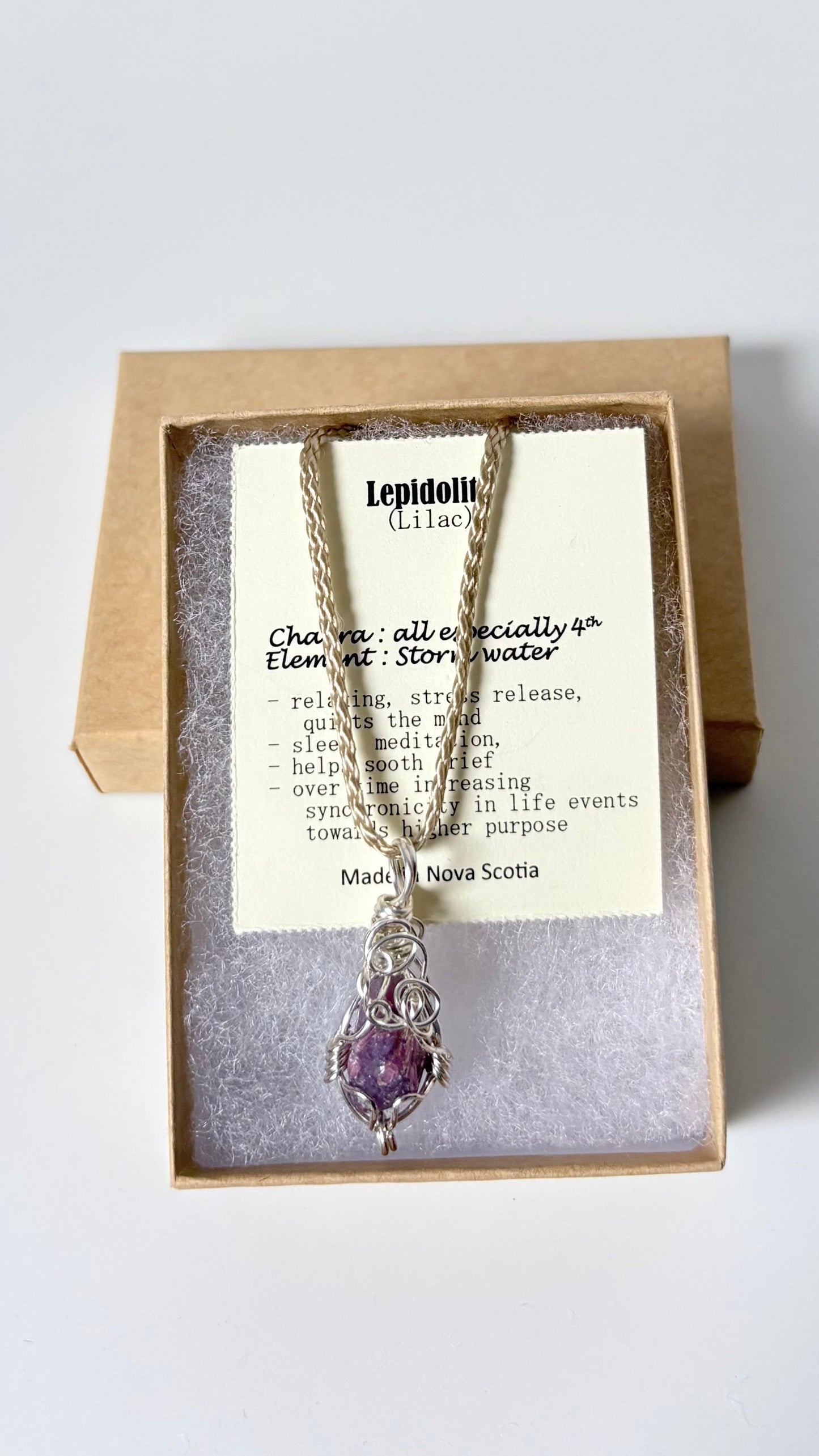 Necklace with a silver wire wrapped lepidolite pendant on an information card with text explaining the folklore and metaphysical properties placed in a brown paper kraft jewelry box for gift giving all on a white surface.