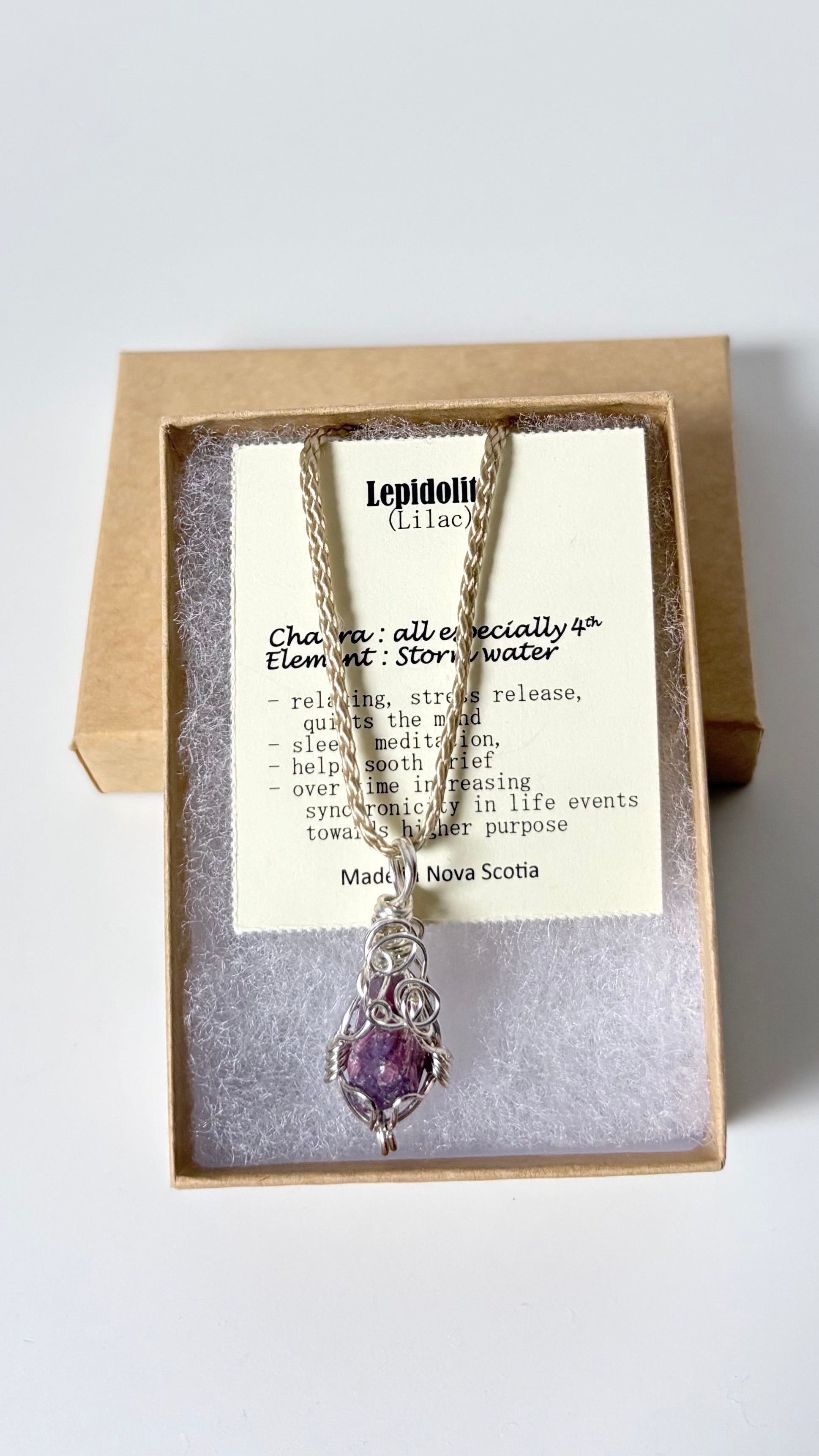 Necklace with a silver wire wrapped lepidolite pendant on an information card with text explaining the folklore and metaphysical properties placed in a brown paper kraft jewelry box for gift giving all on a white surface.