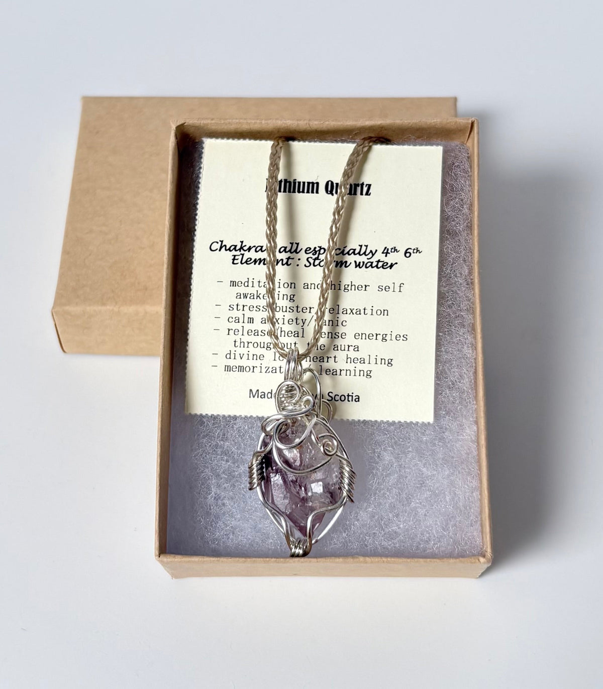 Necklace with a silver wire wrapped lithium quartz point pendant on an information card with text explaining the folklore and metaphysical properties placed in a brown paper kraft jewelry box for gift giving all on a white surface.