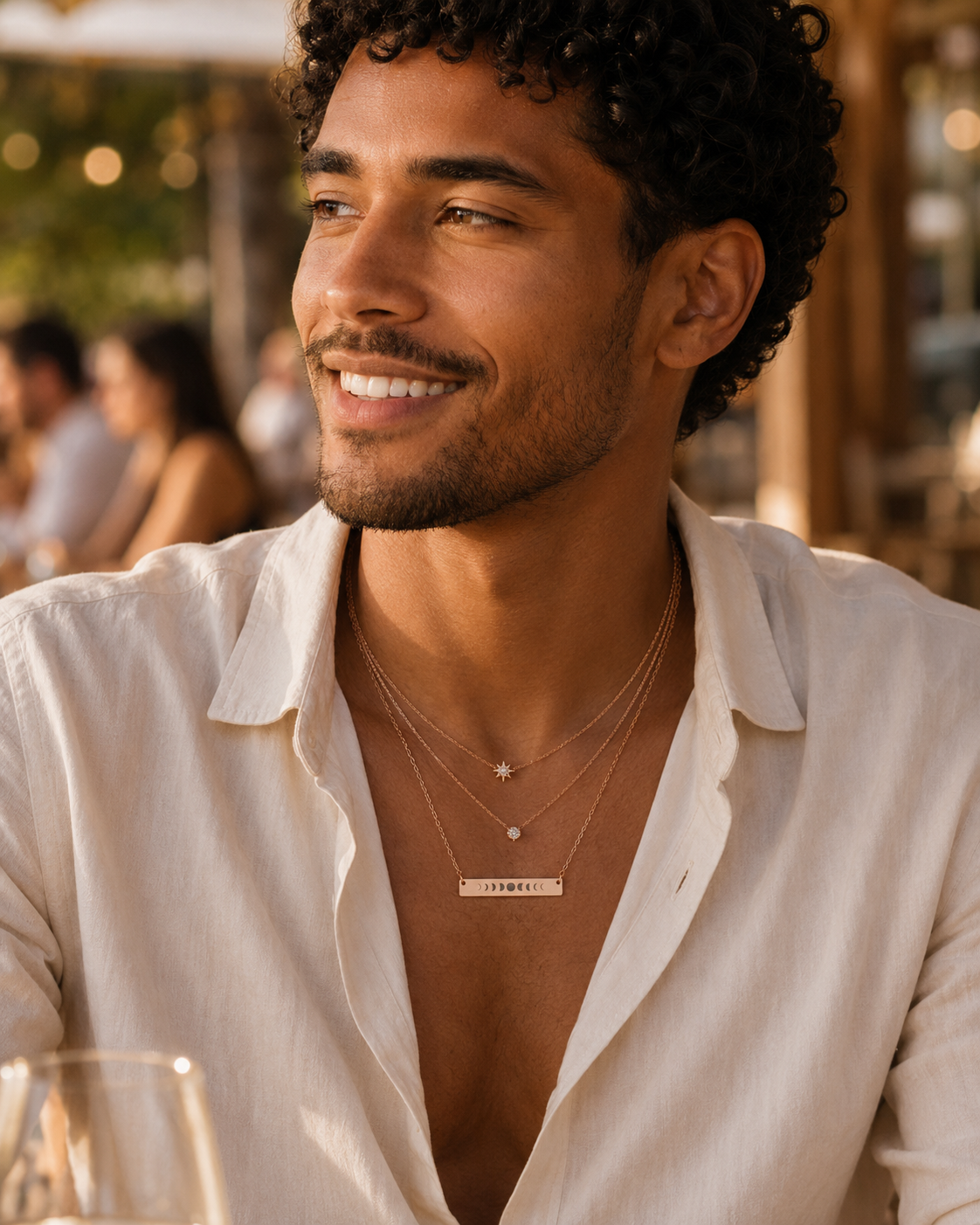 Man wearing a white shirt with layered gold necklaces, sitting outdoors with blurred people in the background. one necklace has a horizontal bar moon phases pendant.