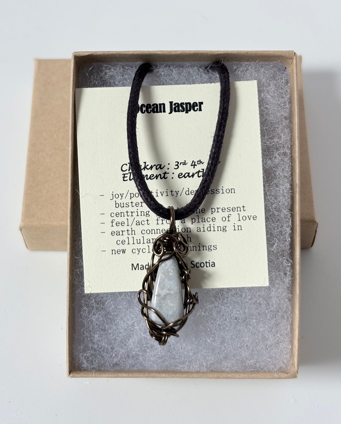 Necklace with an antique brass wire wrapped jasper (ocean) pendant on an information card with text explaining the folklore and metaphysical properties placed in a brown paper kraft jewelry box for gift giving all on a white surface.