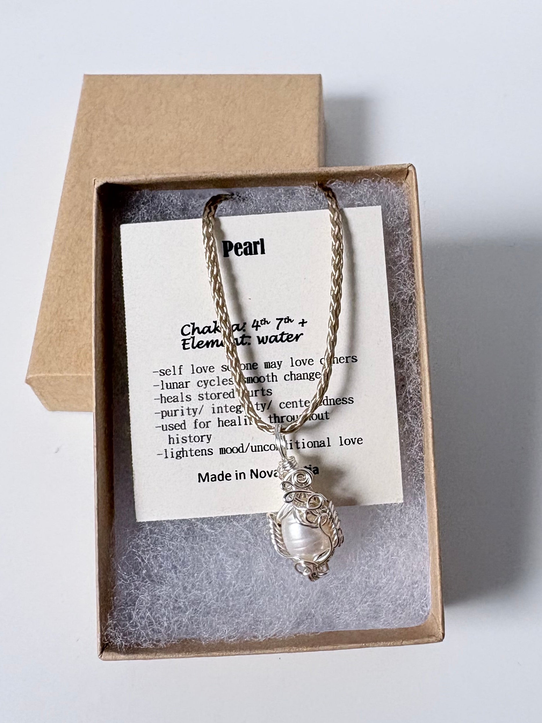 Necklace with a silver wire wrapped pearl pendant on an information card with text explaining the folklore and metaphysical properties placed in a brown paper kraft jewelry box for gift giving all on a white surface.