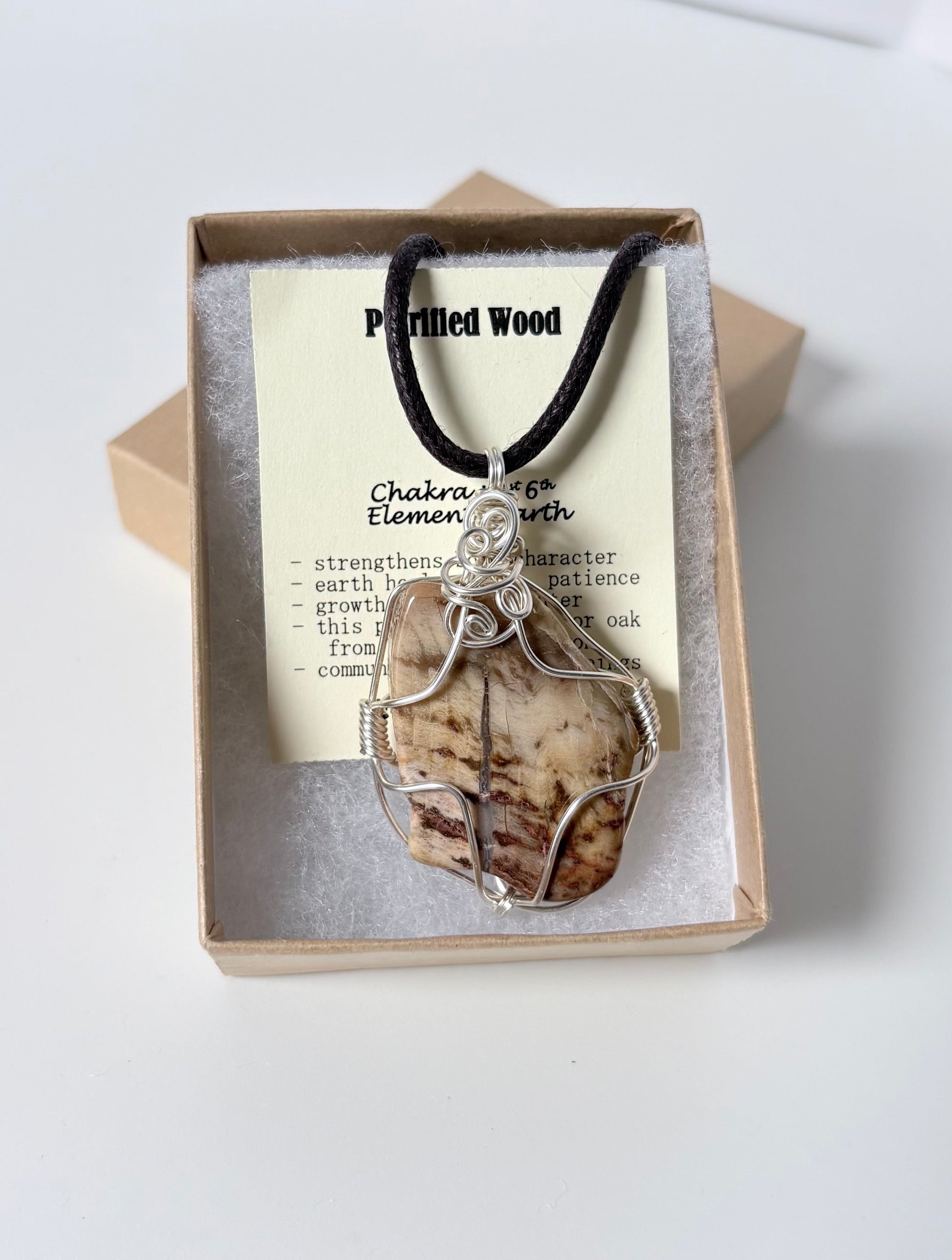Necklace with a silver wire wrapped p mm wood pendant on an information card with text explaining the folklore and metaphysical properties placed in a brown paper kraft jewelry box for gift giving all on a white surface.