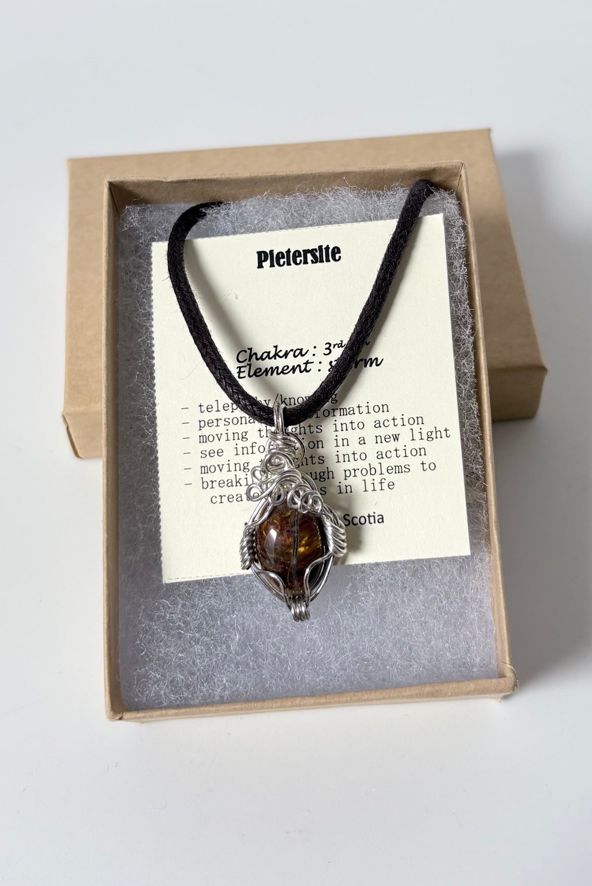Necklace with a silver wire wrapped pietersite pendant on an information card with text explaining the folklore and metaphysical properties placed in a brown paper kraft jewelry box for gift giving all on a white surface.