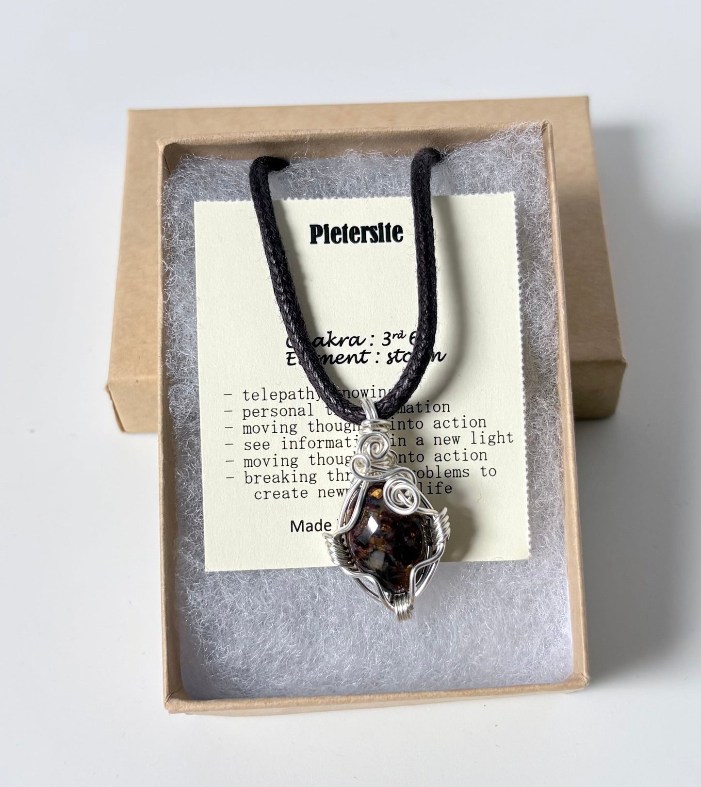 Necklace with a silver wire wrapped Pietersite pendant on an information card with text explaining the folklore and metaphysical properties placed in a brown paper kraft jewelry box for gift giving all on a white surface.