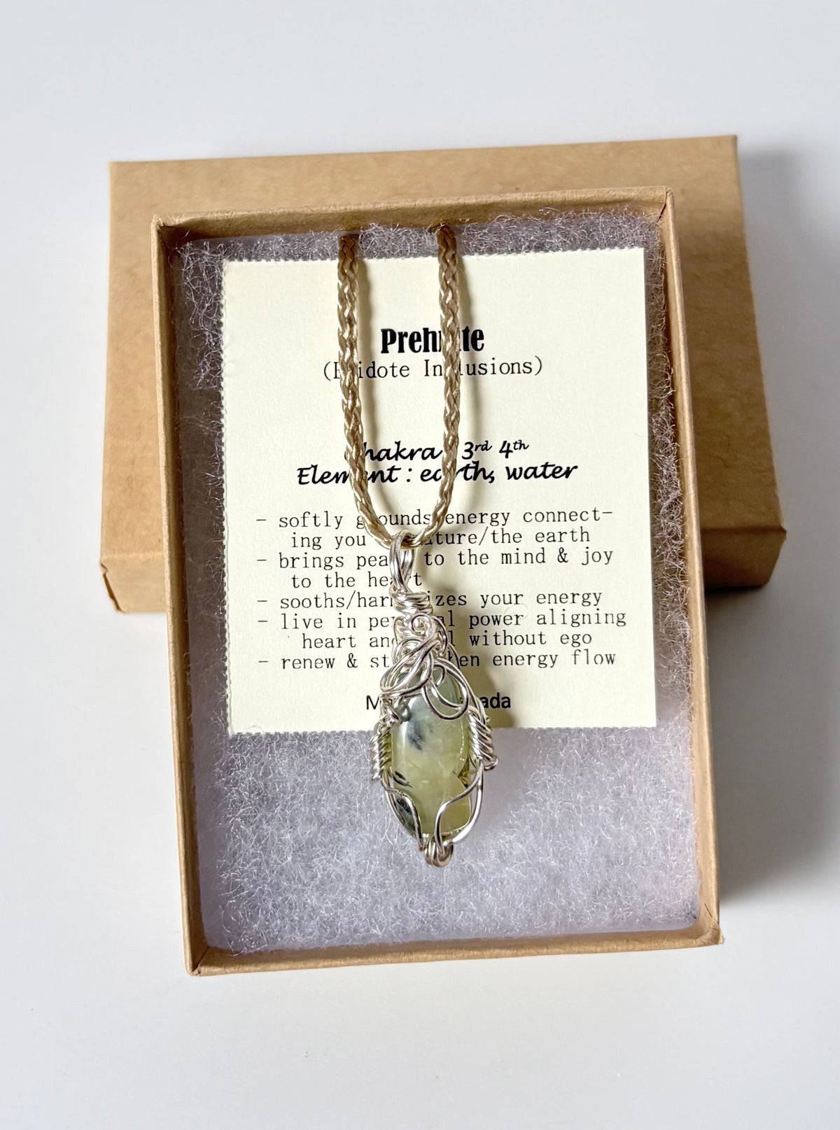 Necklace with a silver wire wrapped Prehnite pendant on an information card with text explaining the folklore and metaphysical properties placed in a brown paper kraft jewelry box for gift giving all on a white surface
